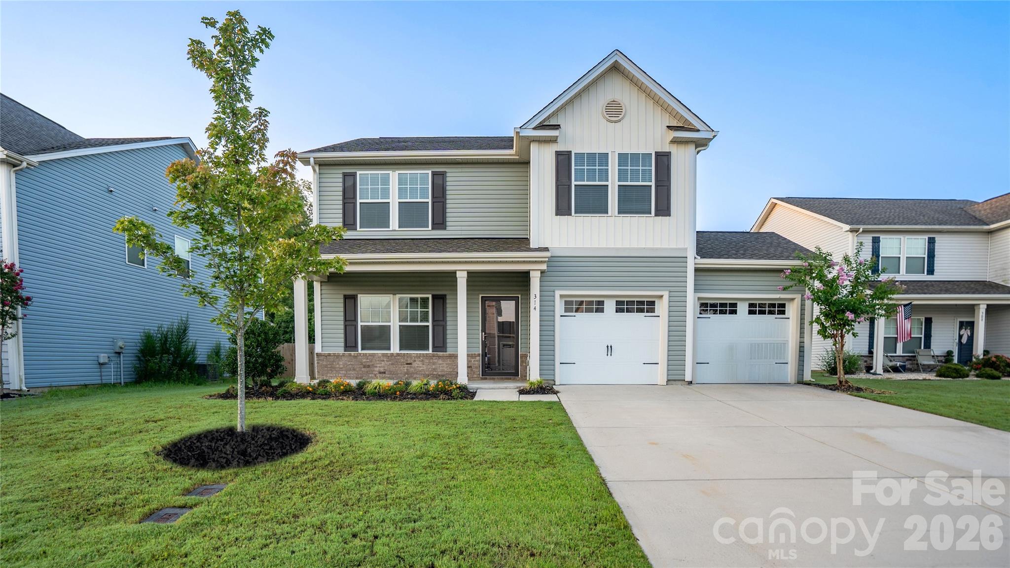 Two-story gray-green home with a covered porch, two white garage doors, and a landscaped front lawn.