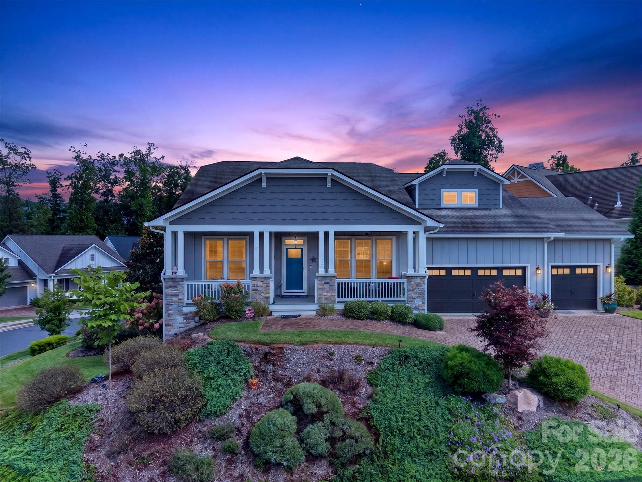 Blue-gray home with a covered porch and two-car garage at dusk, surrounded by landscaped gardens.