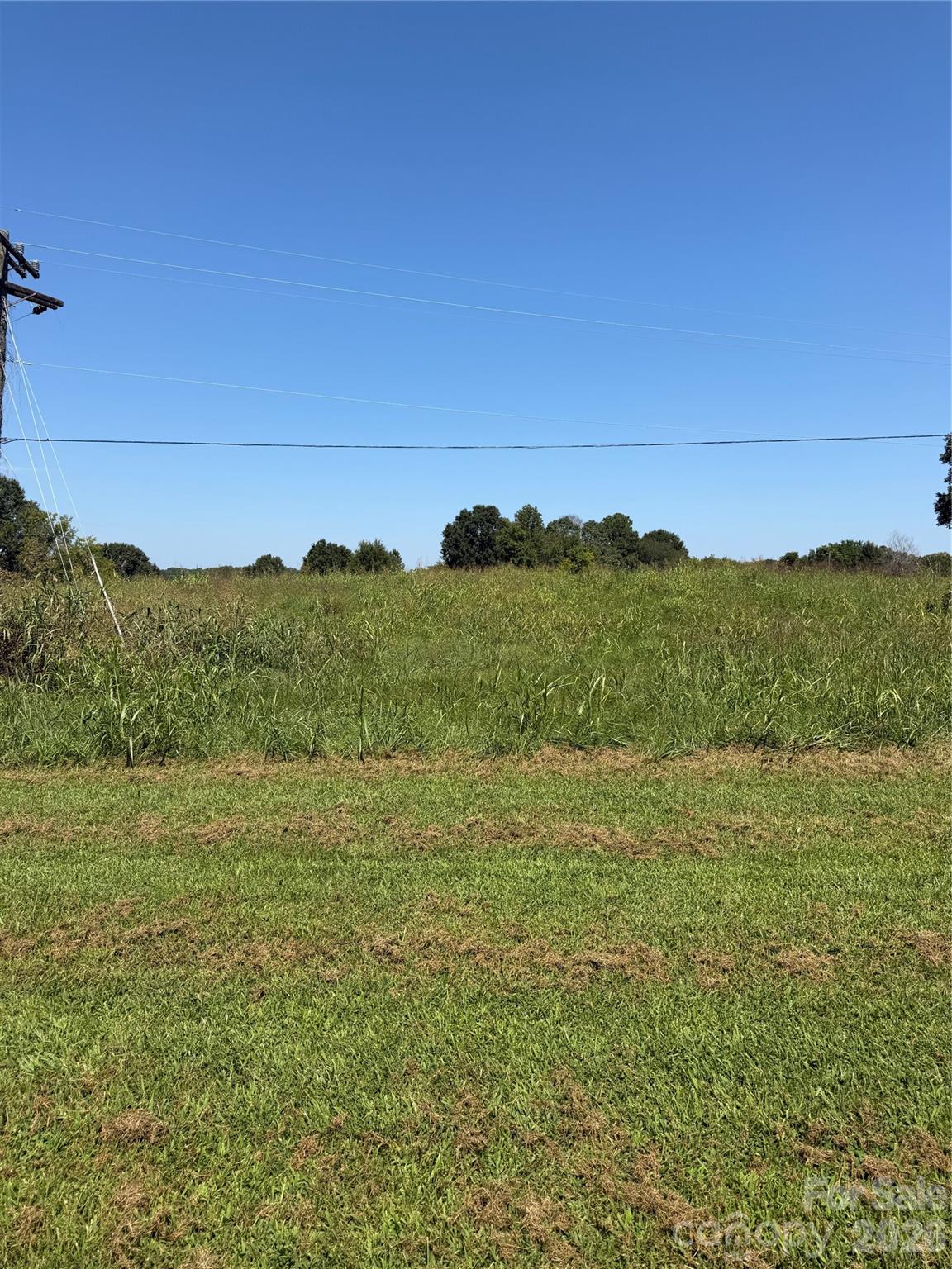 Grassy vacant parcel with tall grass, a mown foreground, distant trees, and overhead utility lines.