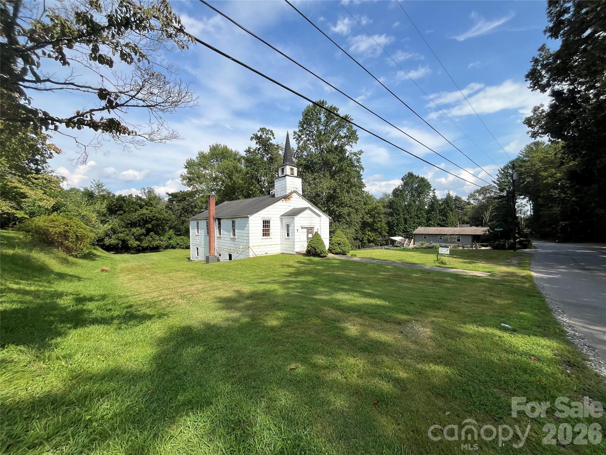 White church with a tall steeple on a grassy lot beside a tree-lined road.