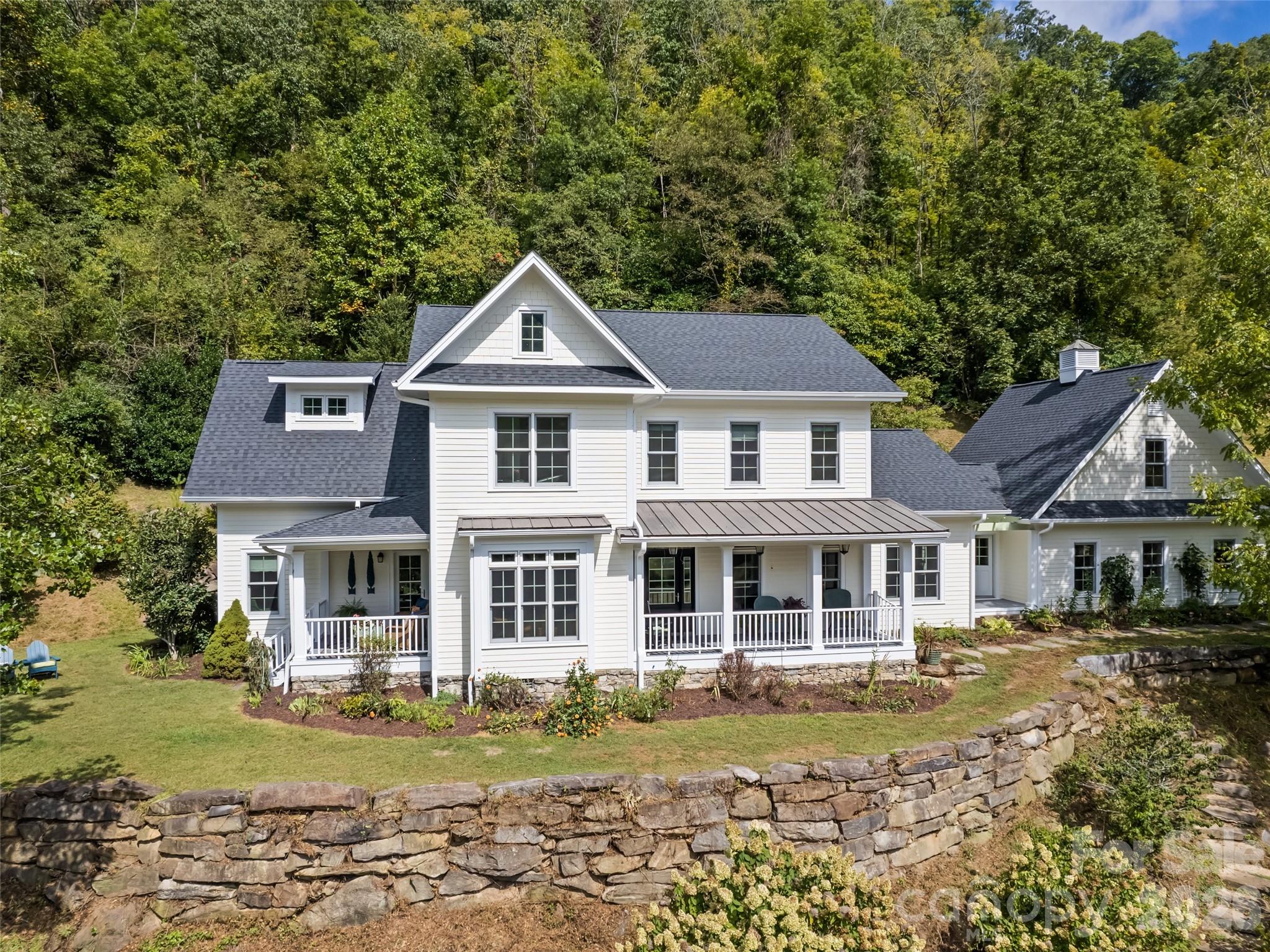 White two-story house with covered porches, wooded hillside, and curved stone retaining wall.
