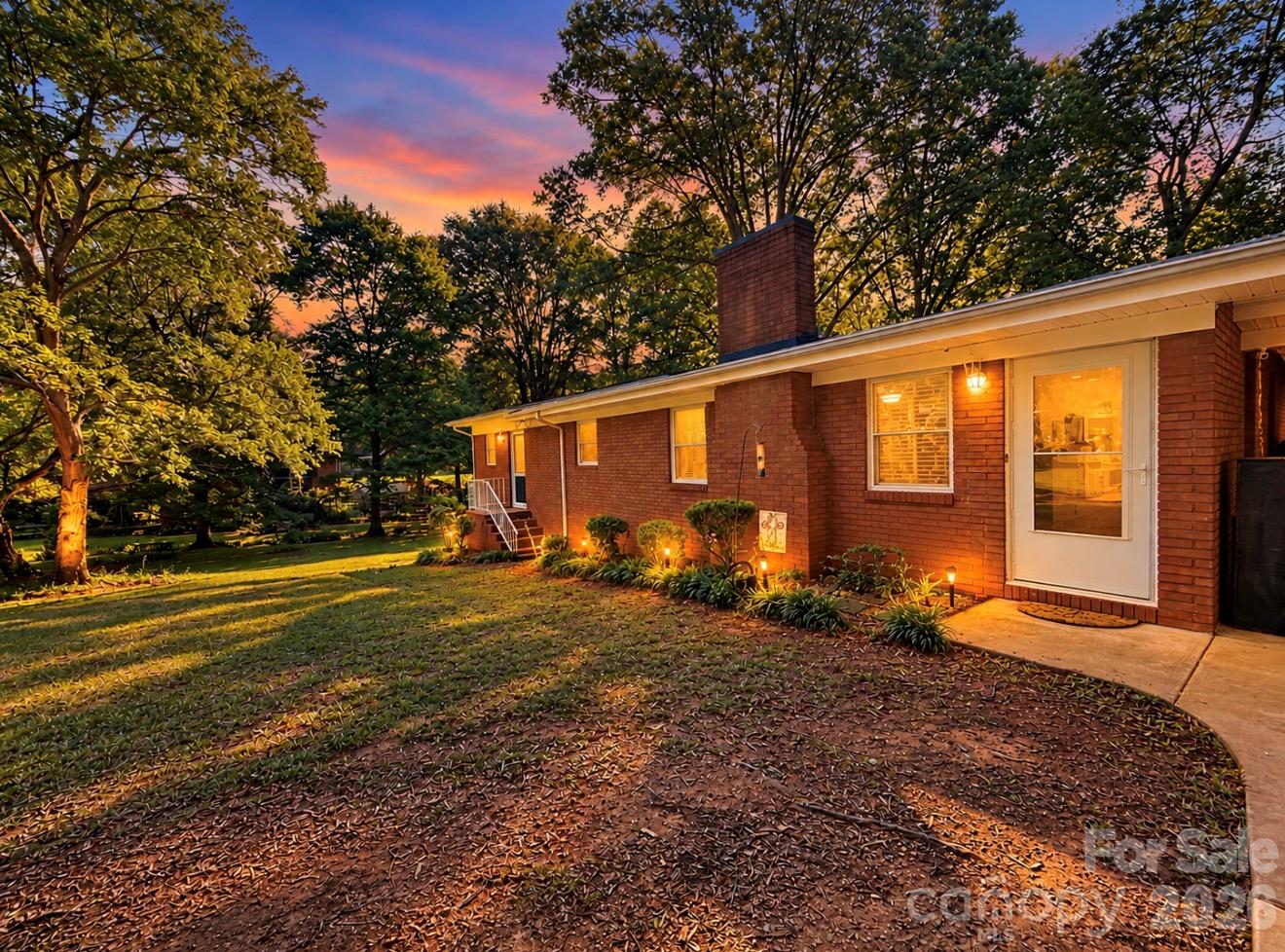 Red-brick ranch home at dusk, with warmly lit windows and mature trees framing the landscaped yard.