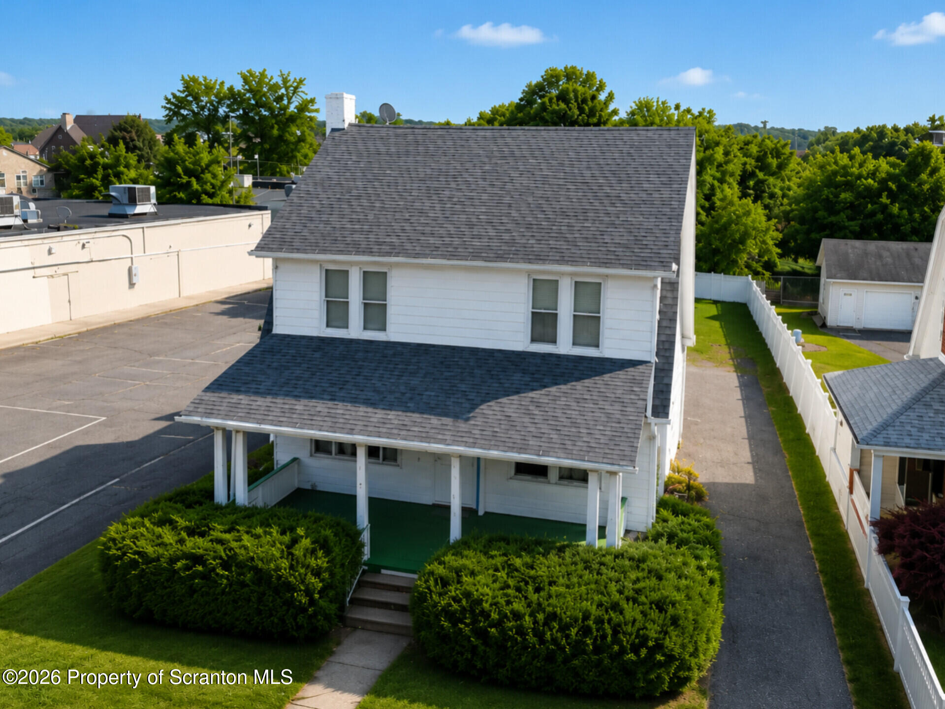 White two-story house with gray shingle roofs, a covered porch, and trimmed front shrubs.