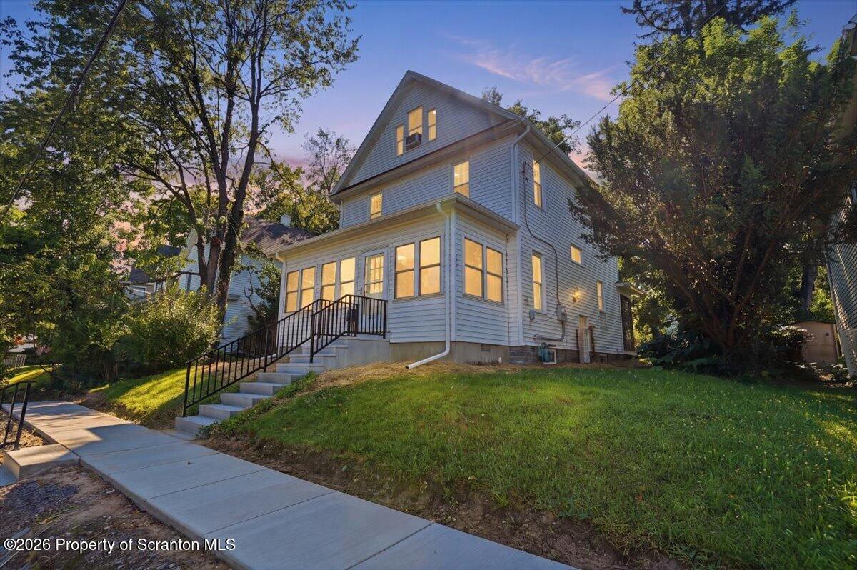 Two-story gray house with a brightly lit enclosed porch, concrete steps, and a grassy front slope.