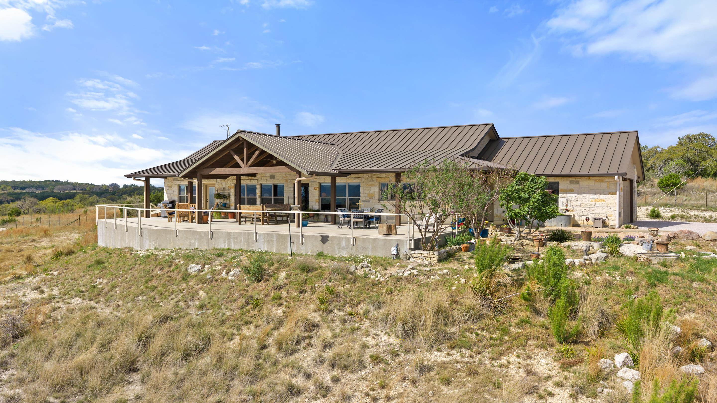 Stone home with a brown metal roof and covered patio on a rocky, grassy hillside.