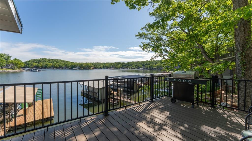 Lakeside deck overlooking a calm lake and wooded shoreline, with docks and a barbecue grill.