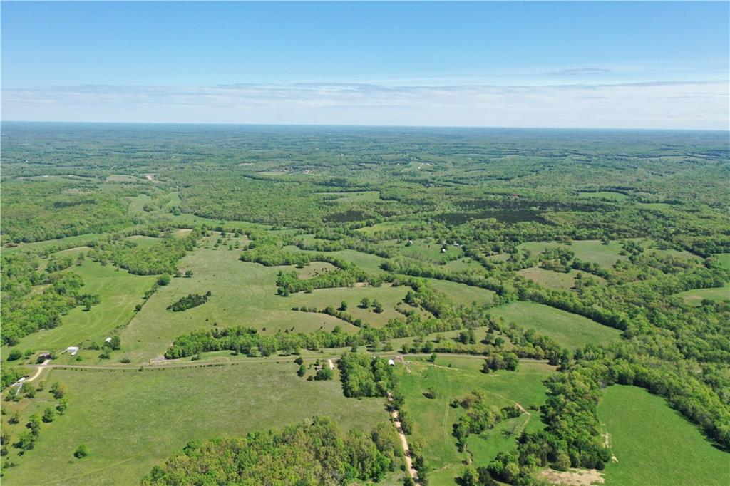 Aerial view of green fields, wooded patches, and scattered rural homes beneath a broad blue sky.