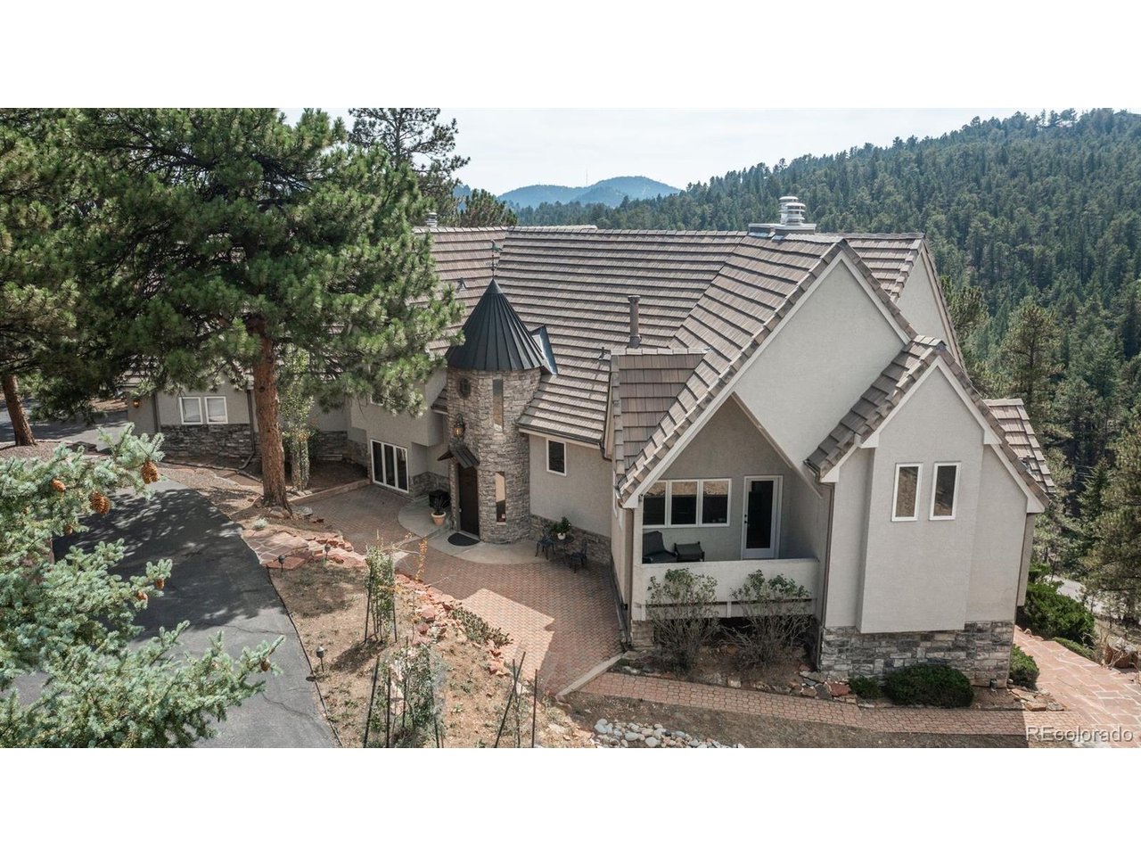 Mountain home with a stone turret and tiled roof among pine trees and forested hills.