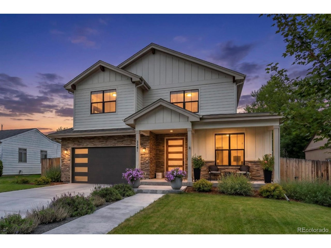 Two-story white house with stone accents, glowing windows, and a landscaped front yard at dusk.