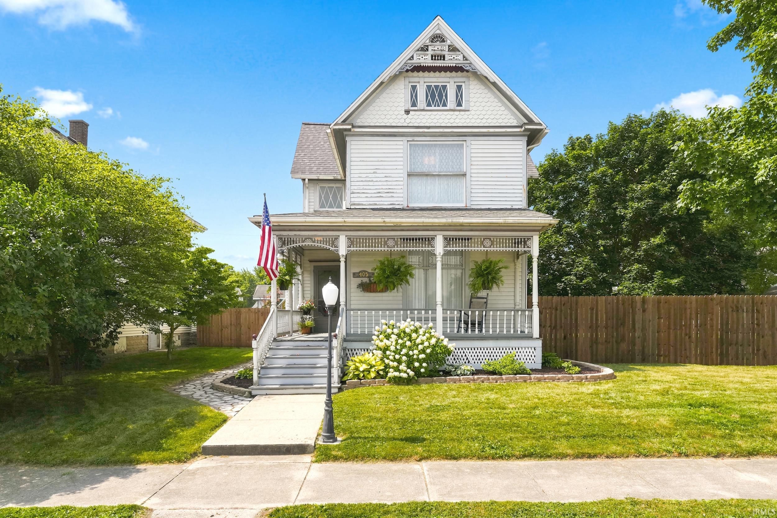 White Victorian house with a steep gable, ornate porch, American flag, and flower garden.