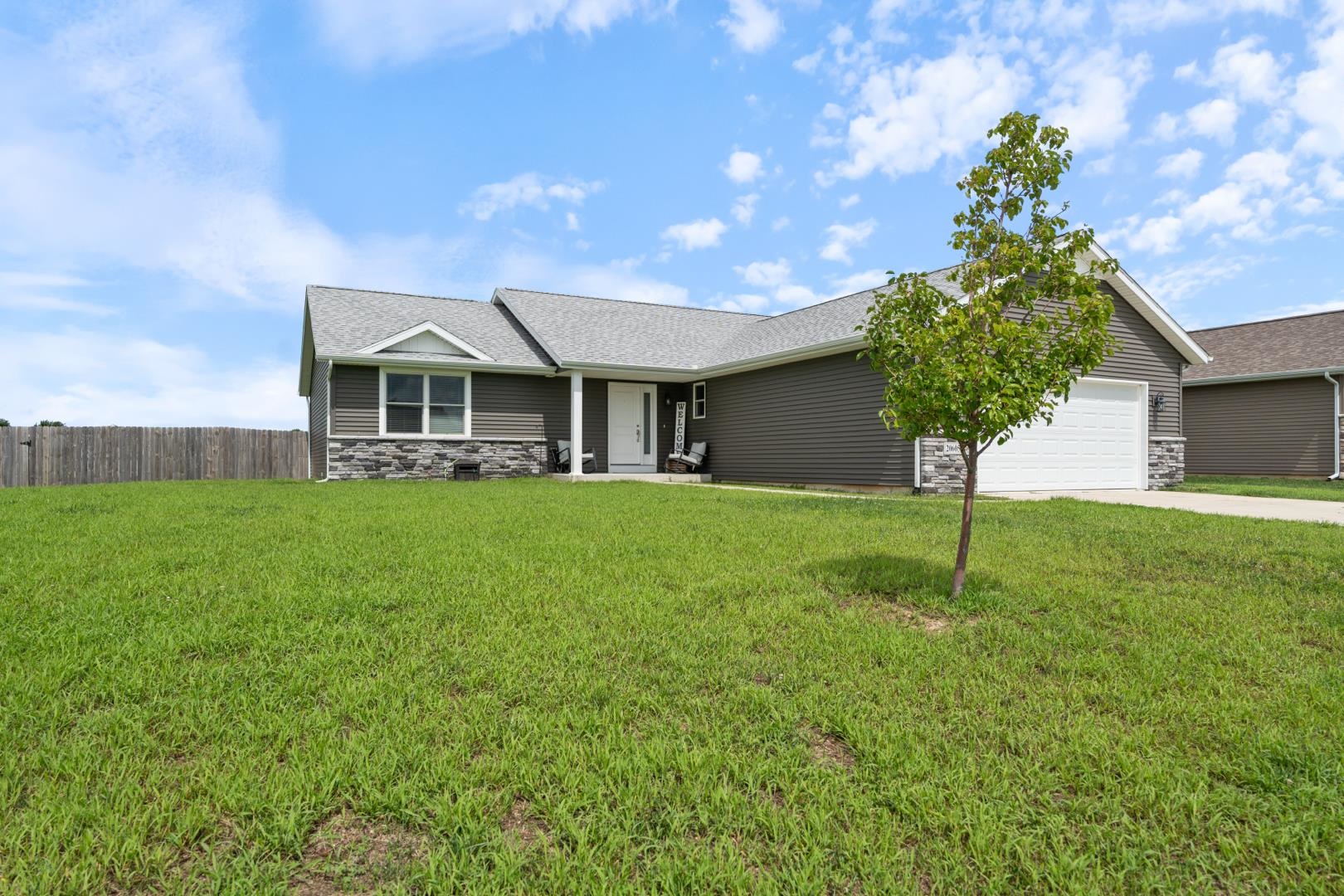 Gray single-story house with stone accents, a white garage, covered porch, and broad green lawn.
