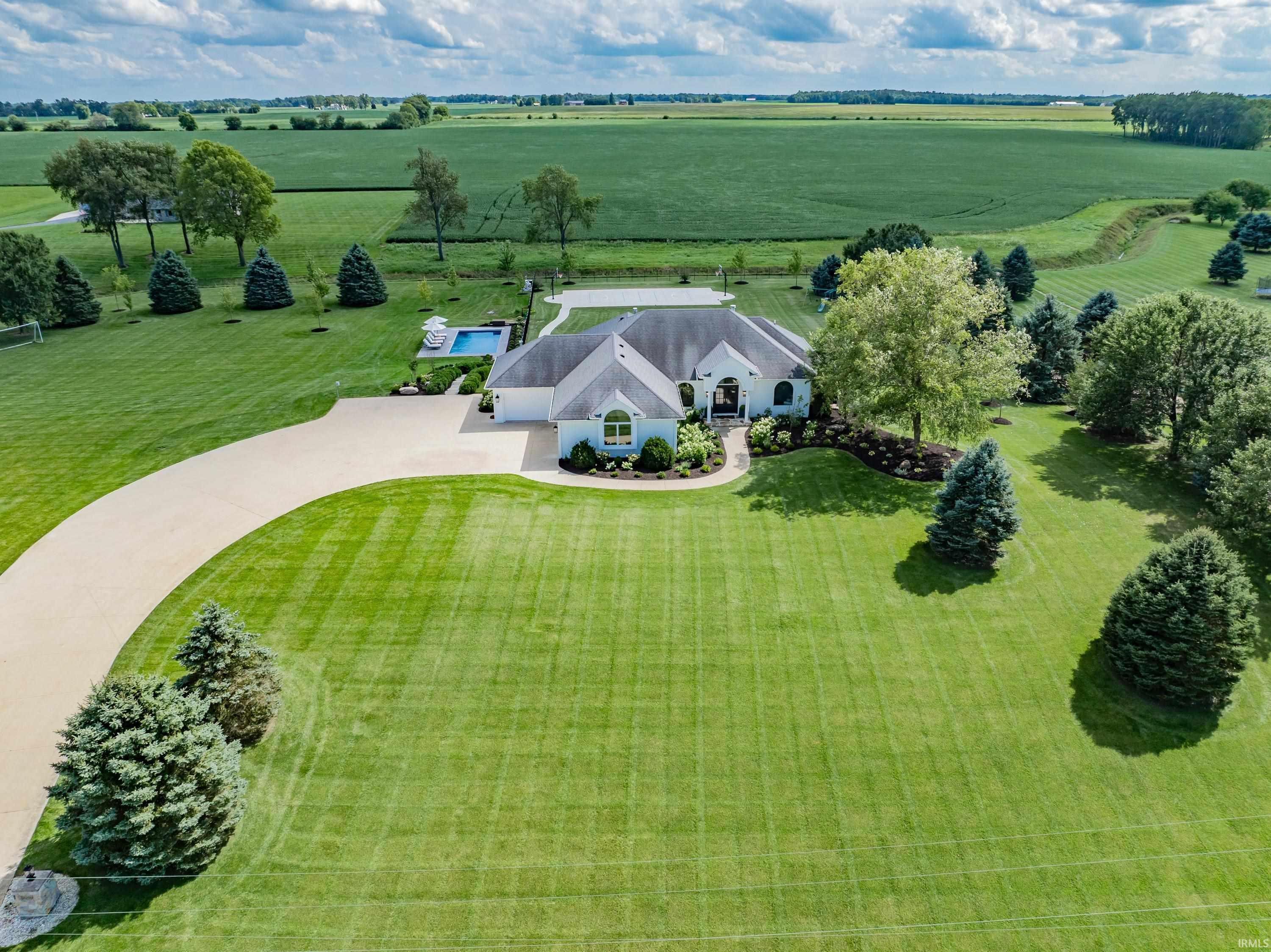Aerial view of a white home with a curved driveway, pool, and expansive lawn bordered by farmland.