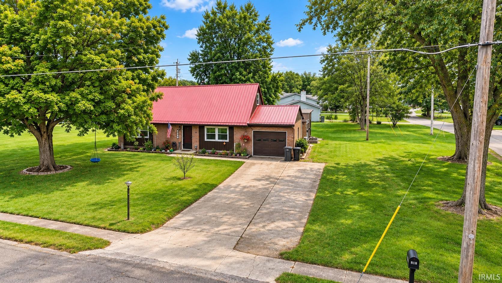 Brick home with a red metal roof and attached garage, set behind a broad driveway and mature trees.