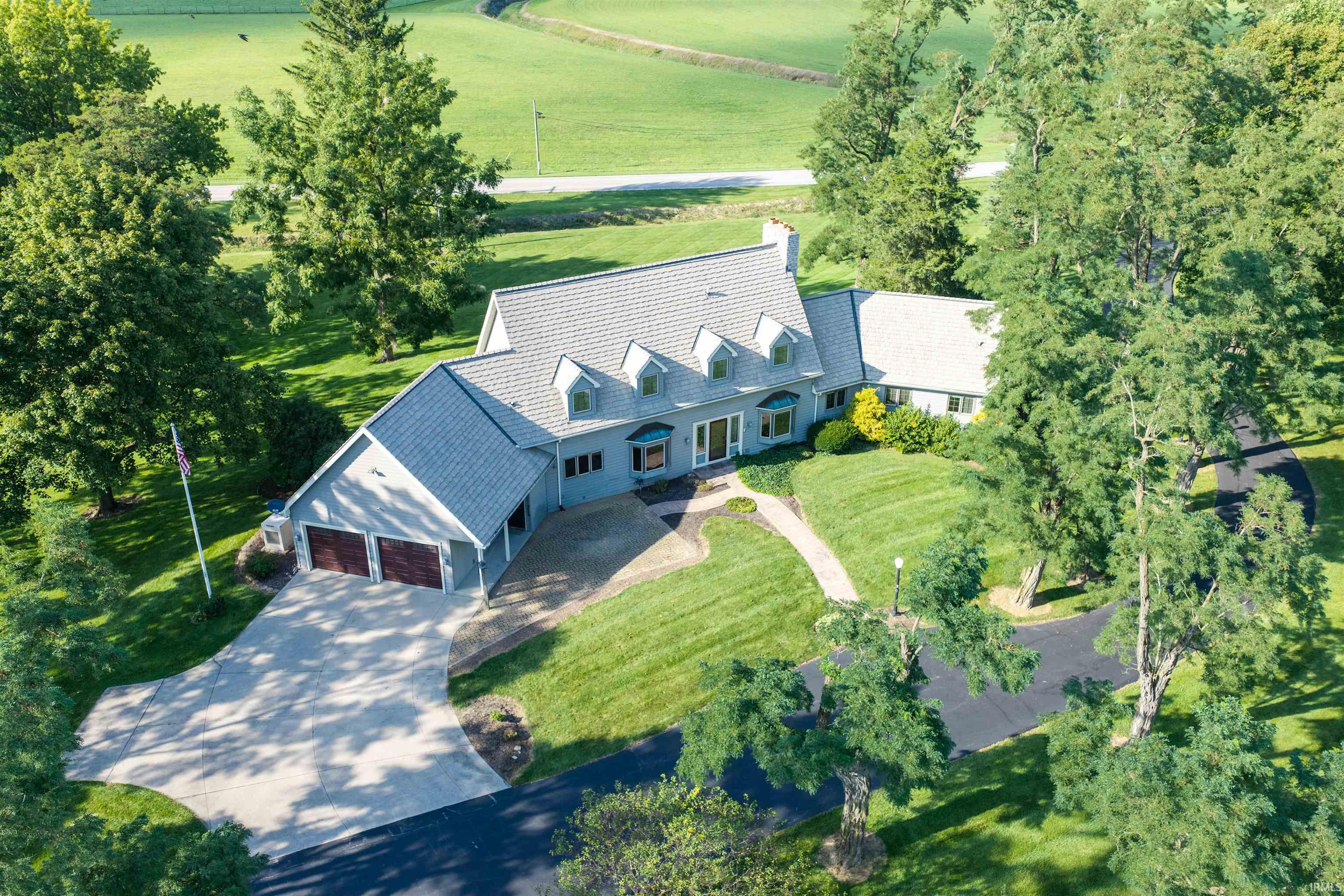 Aerial view of a gray-blue home with multiple dormers, a two-car garage, and a tree-lined lawn.