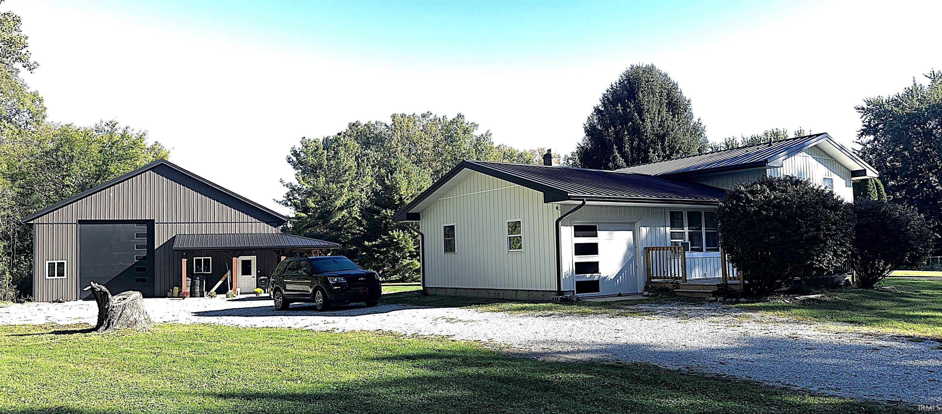 Light-colored metal-sided home with a dark roof, detached garage, and gravel driveway.
