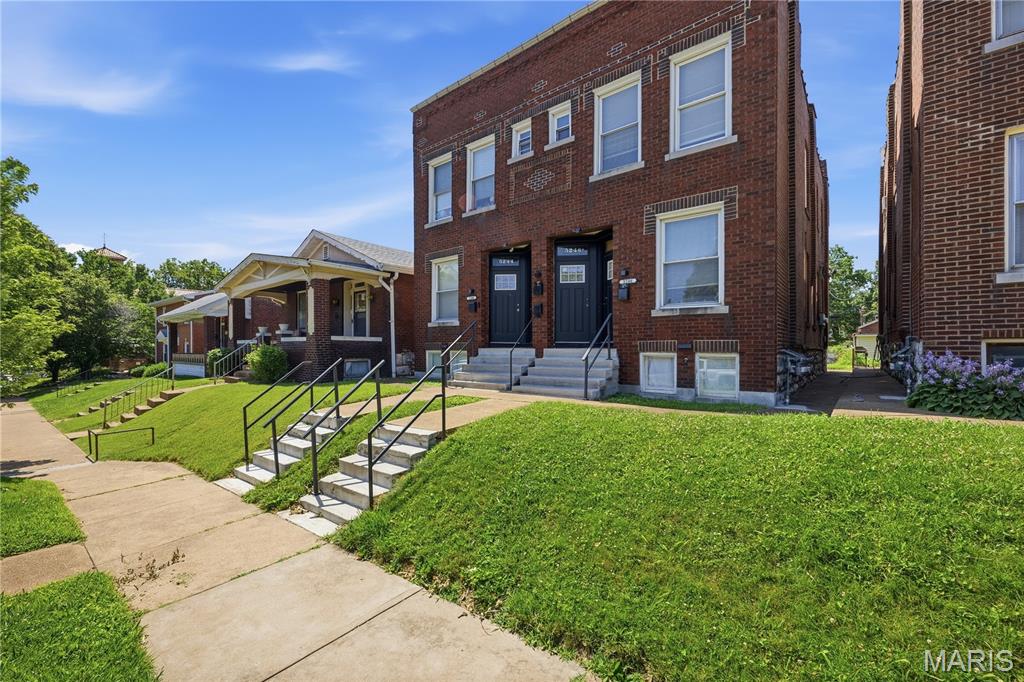 Red-brick multi-unit building with twin entrances, front steps, black railings, and a green lawn.