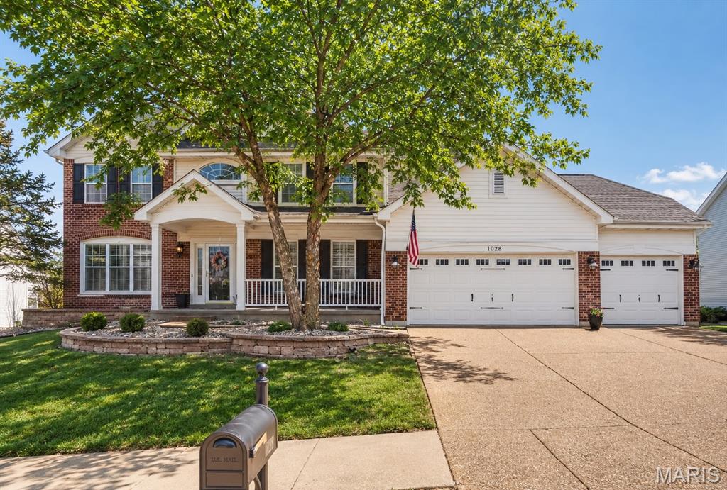 Two-story brick-and-siding home with a covered porch, three-car garage, mature front-yard tree, and American flag.