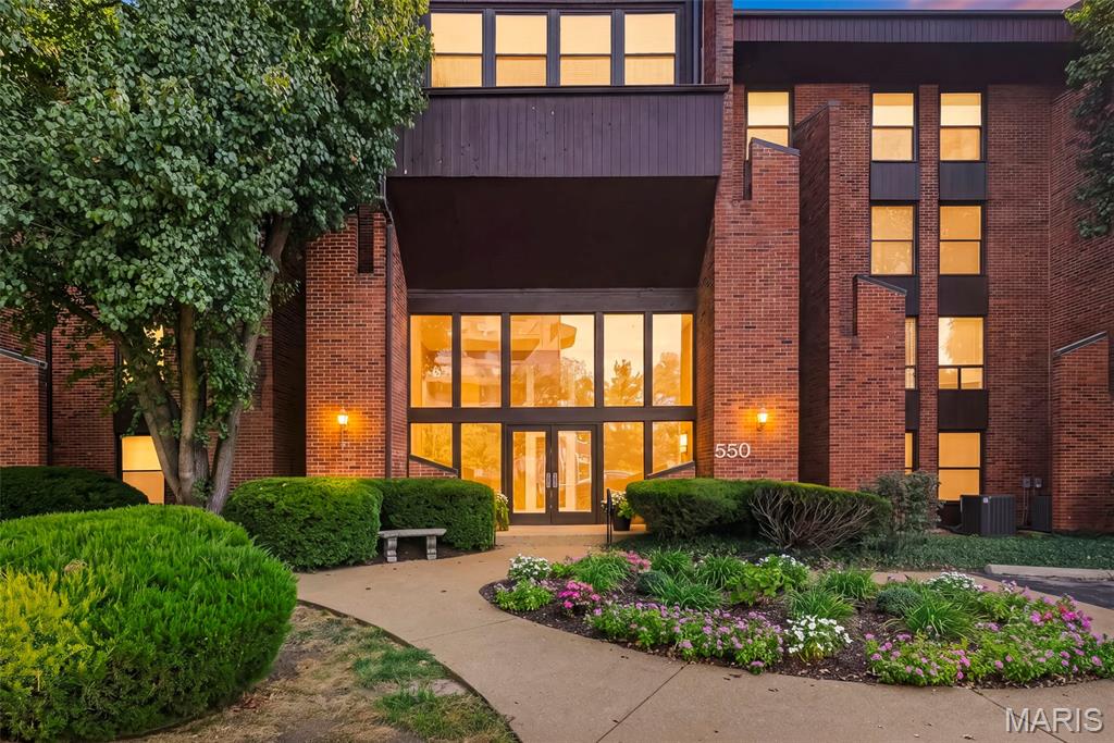 Red-brick residential building with a recessed glass entrance, glowing windows, and landscaped gardens at dusk.