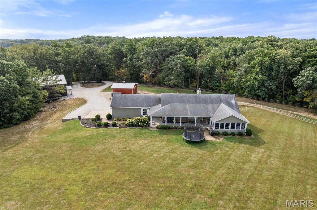 Aerial view of a gray-roofed home with a red outbuilding, trampoline, and wooded property.