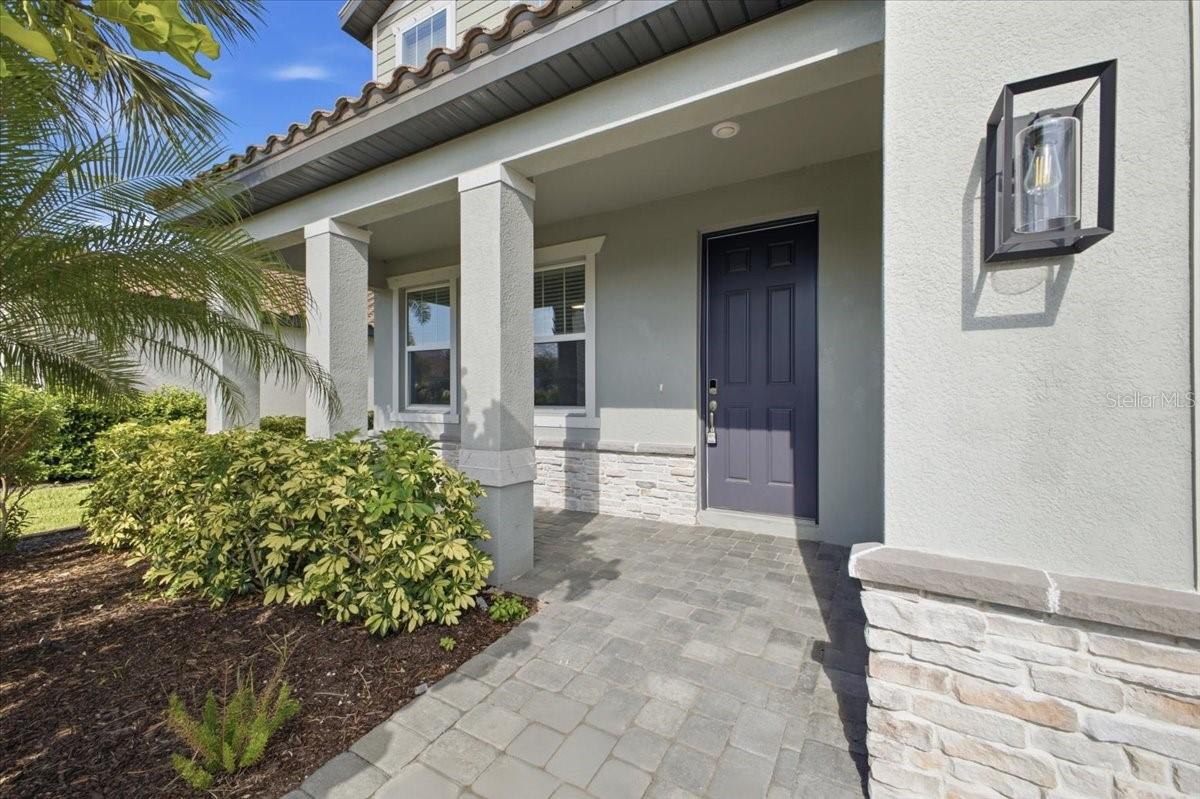Home entrance with a covered porch, dark blue door, stone accents, and tropical landscaping.