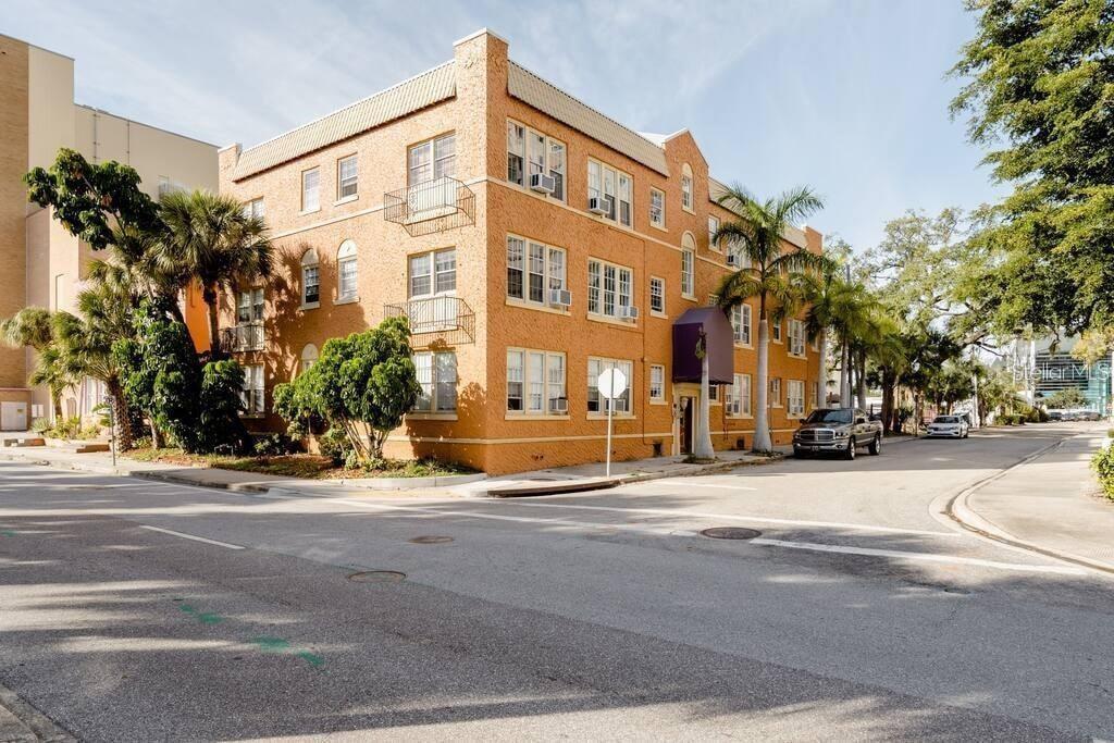 Three-story orange brick building on a corner, with a purple entrance awning and palm trees.
