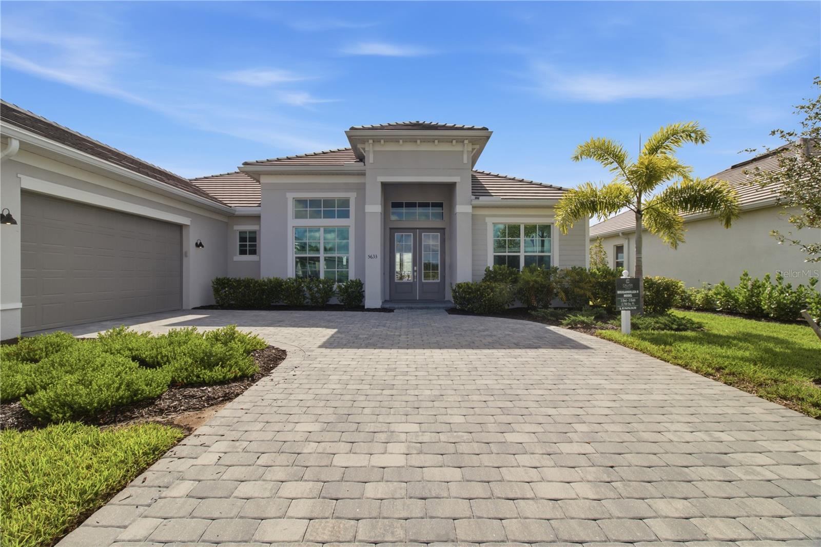 Modern gray single-story home with a two-car garage, wide paver driveway, and palm trees.