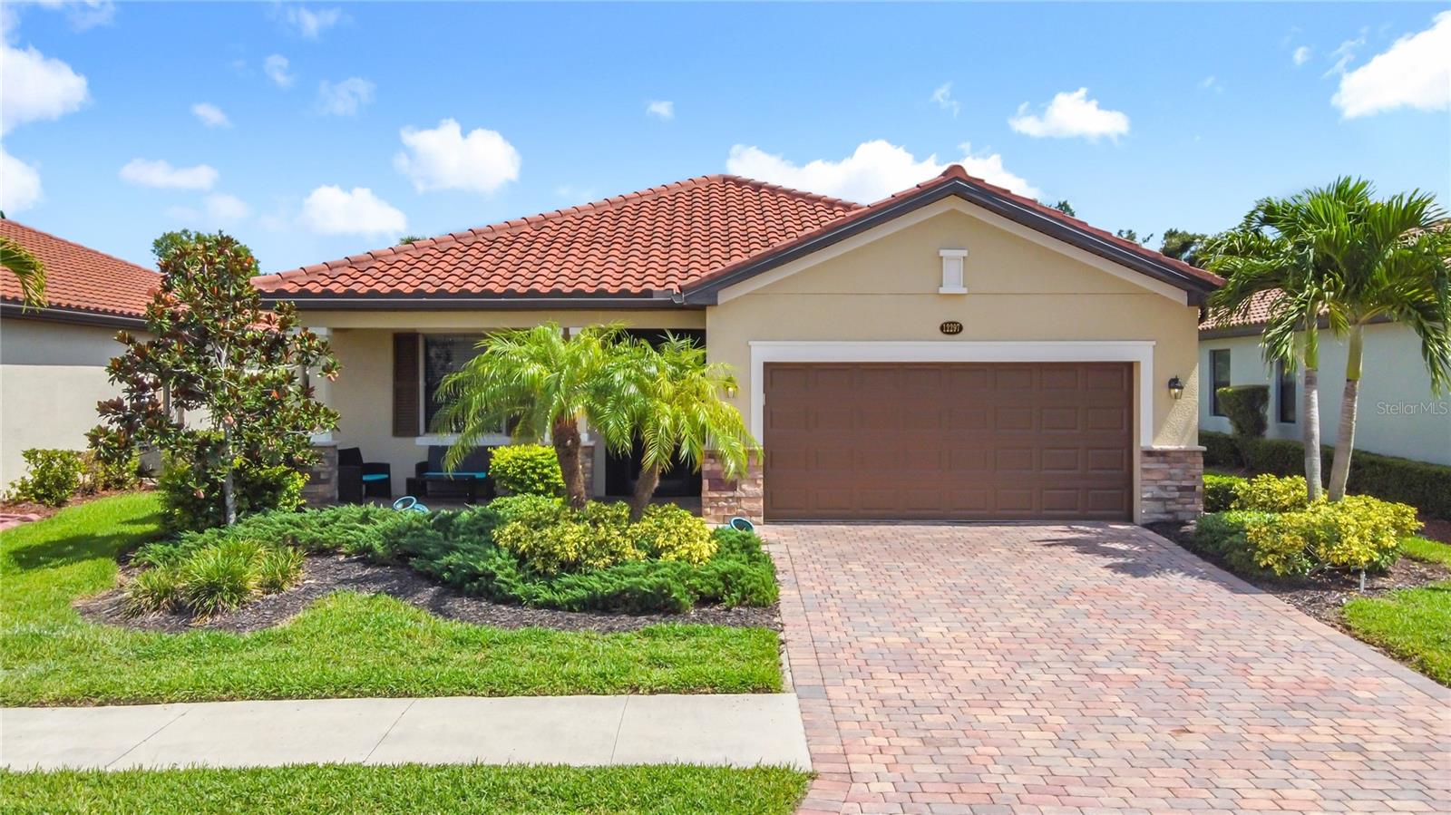 Single-story stucco home with a terracotta tile roof, two-car garage, paver driveway, and tropical landscaping.