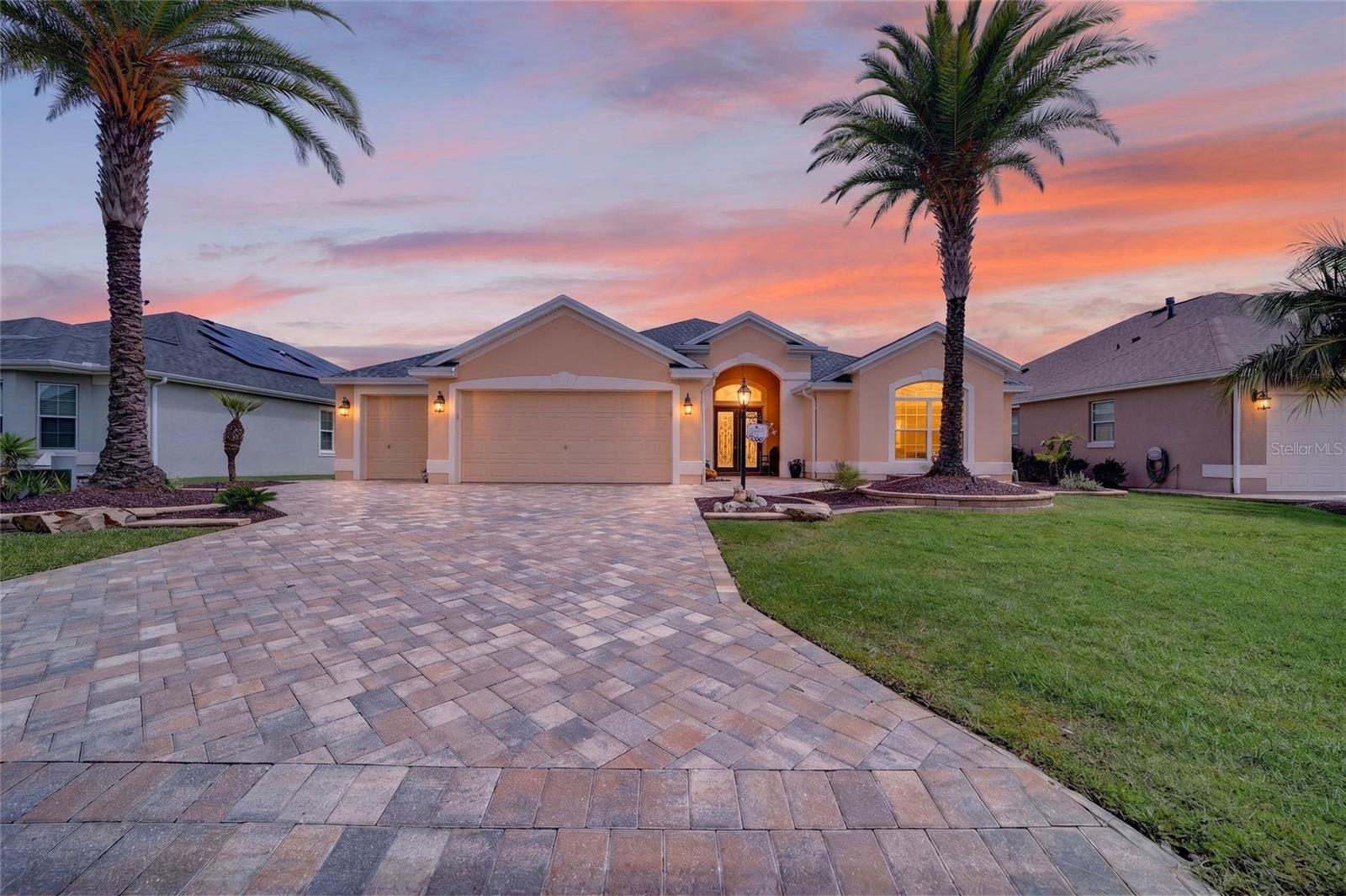 Peach single-story home with a paver driveway, palm trees, and warmly lit entry at dusk.
