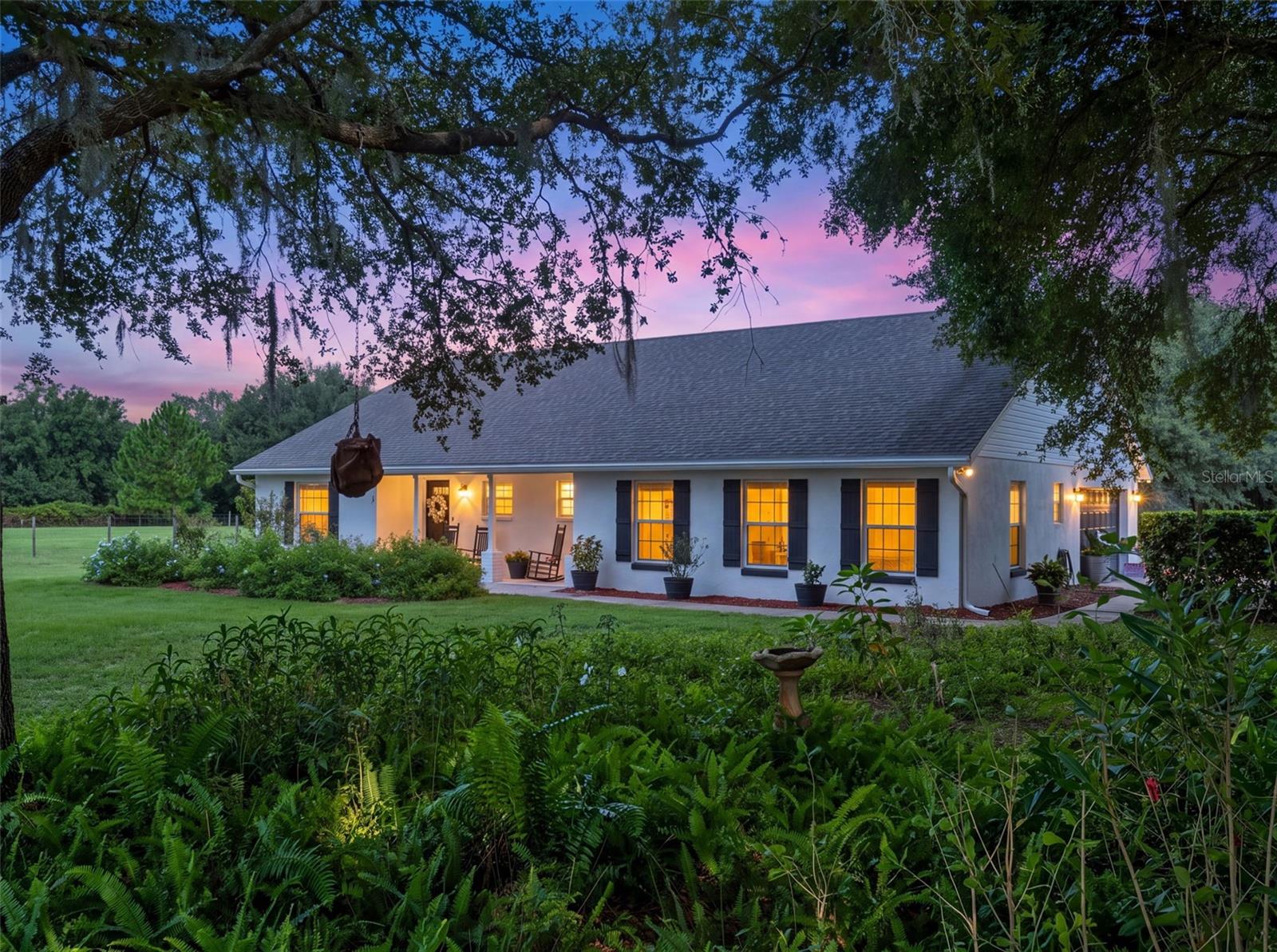 White single-story house glowing at dusk beneath sprawling oak branches, surrounded by a fern-filled garden.