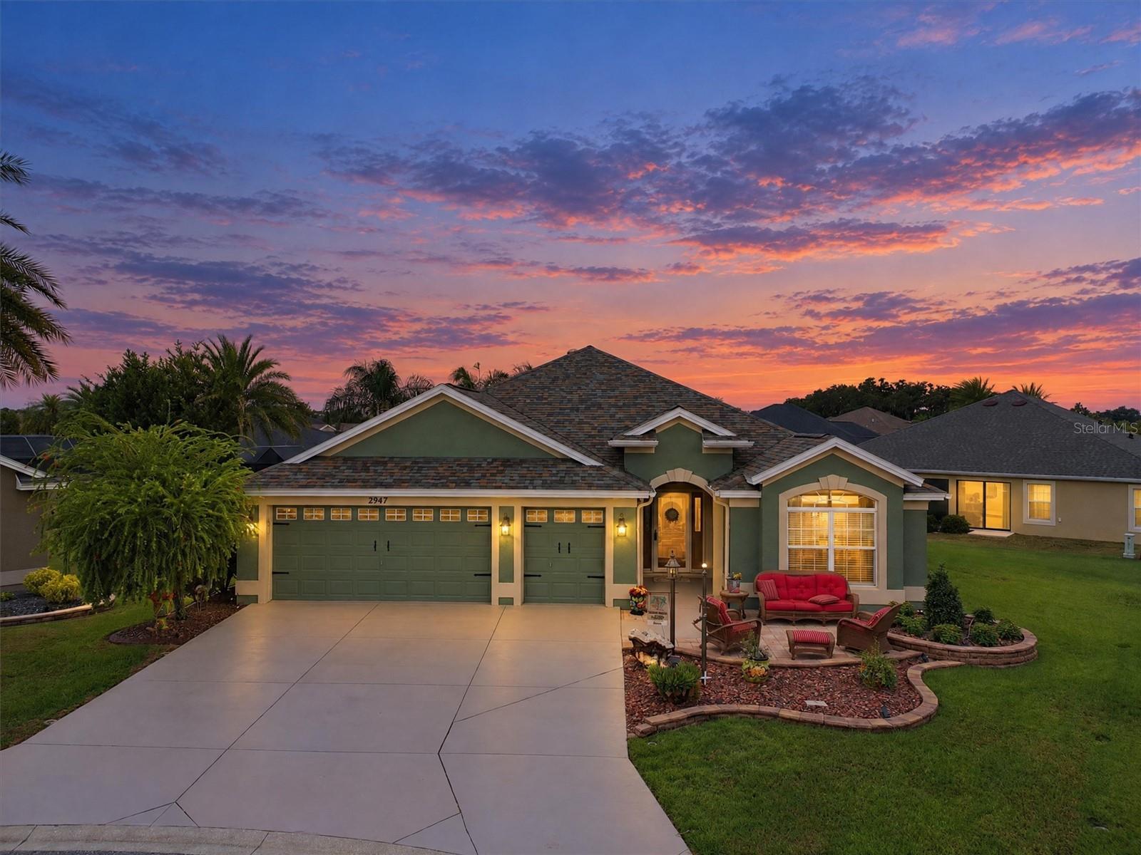 Green stucco home with a wide driveway and outdoor seating beneath a colorful dusk sky.