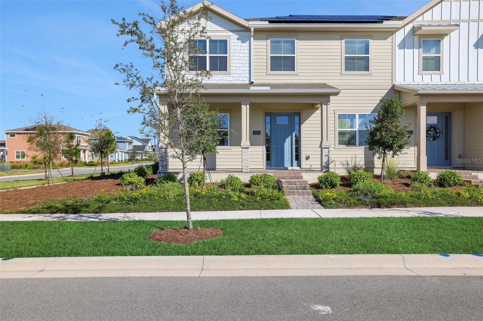 Two-story beige townhome with blue doors, rooftop solar panels, and landscaped front beds.