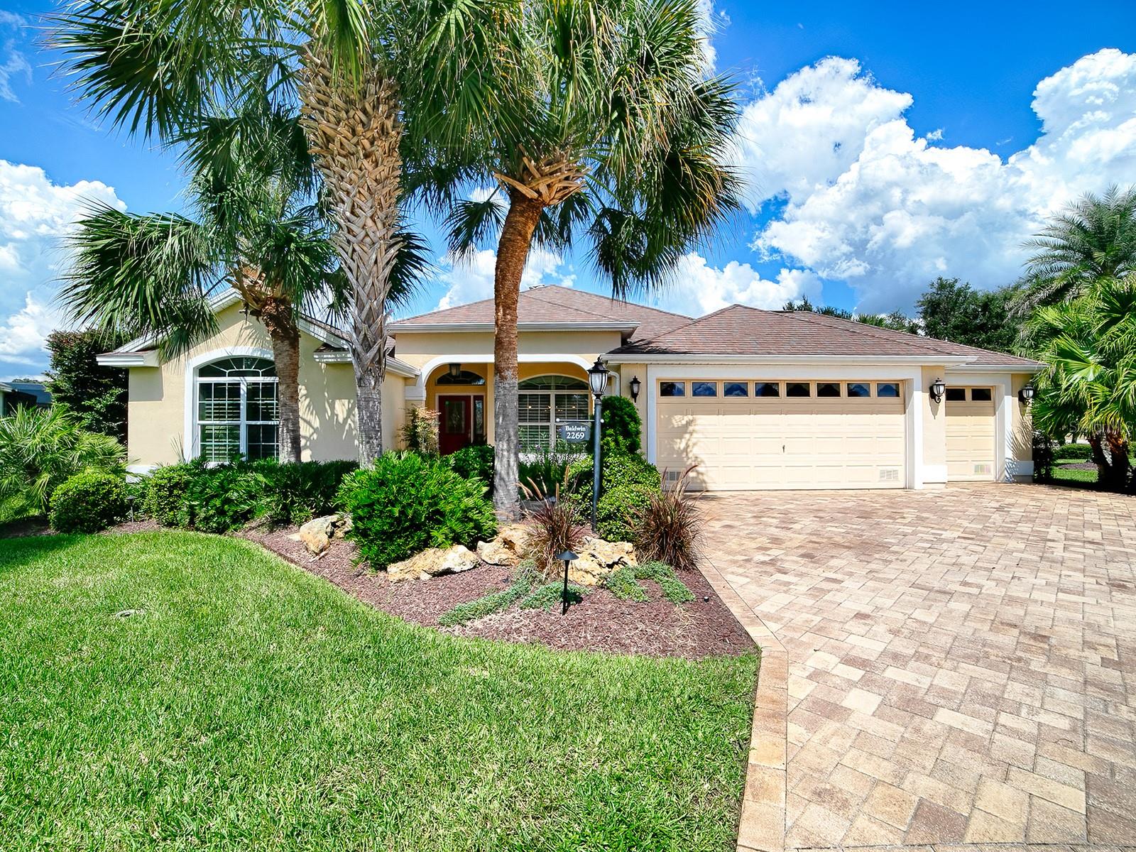 Single-story tan home with a three-car garage, paver driveway, and palm trees.