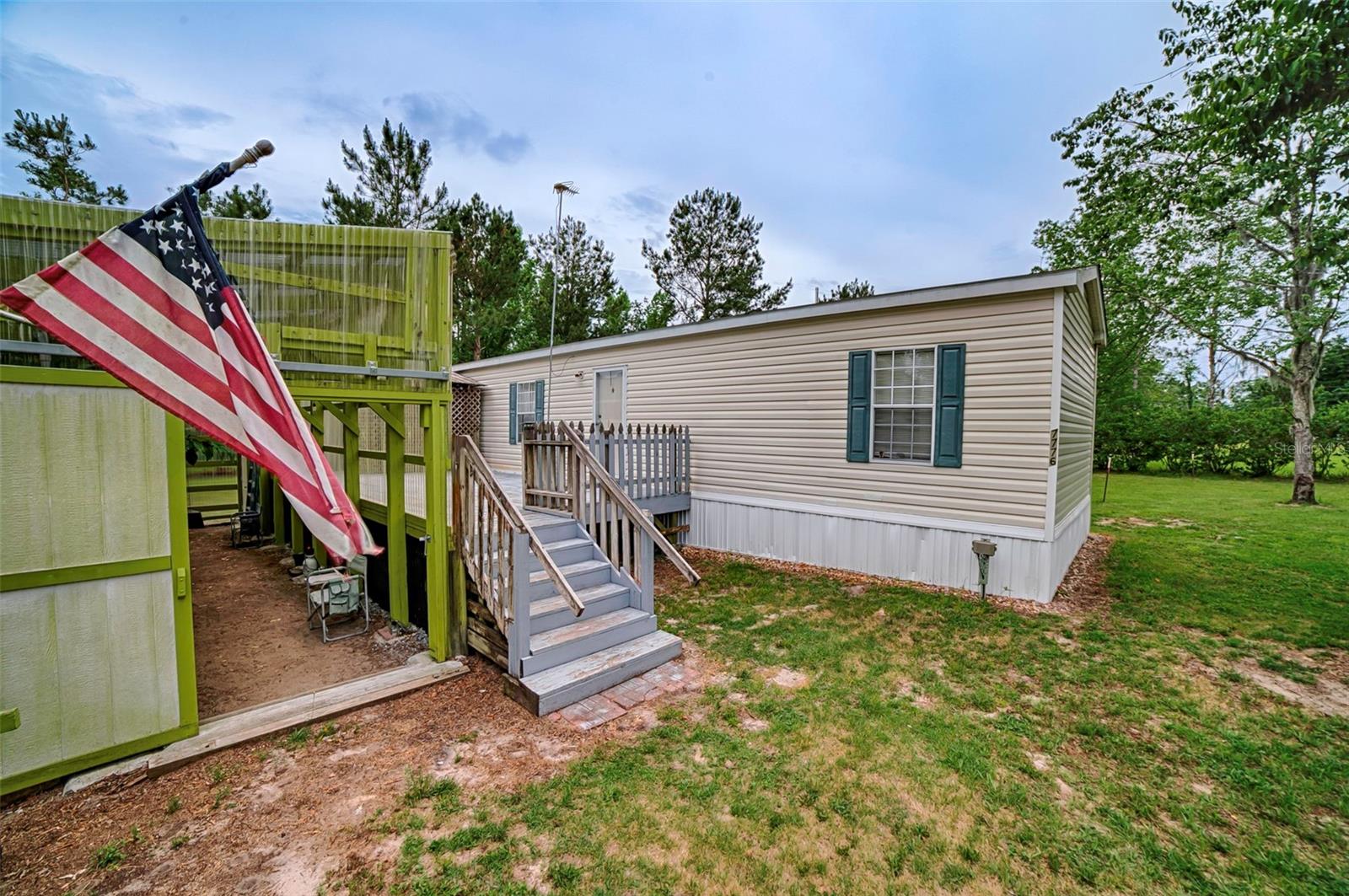 Beige manufactured home with teal shutters, raised wooden deck, stairs, and an American flag.