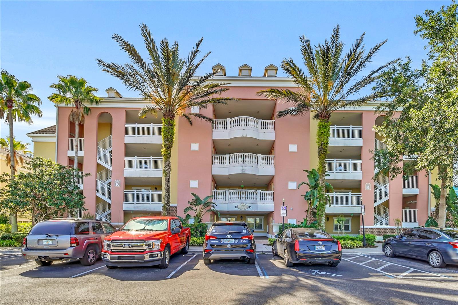 Condominium building with white balconies and palm trees above a row of parked cars.