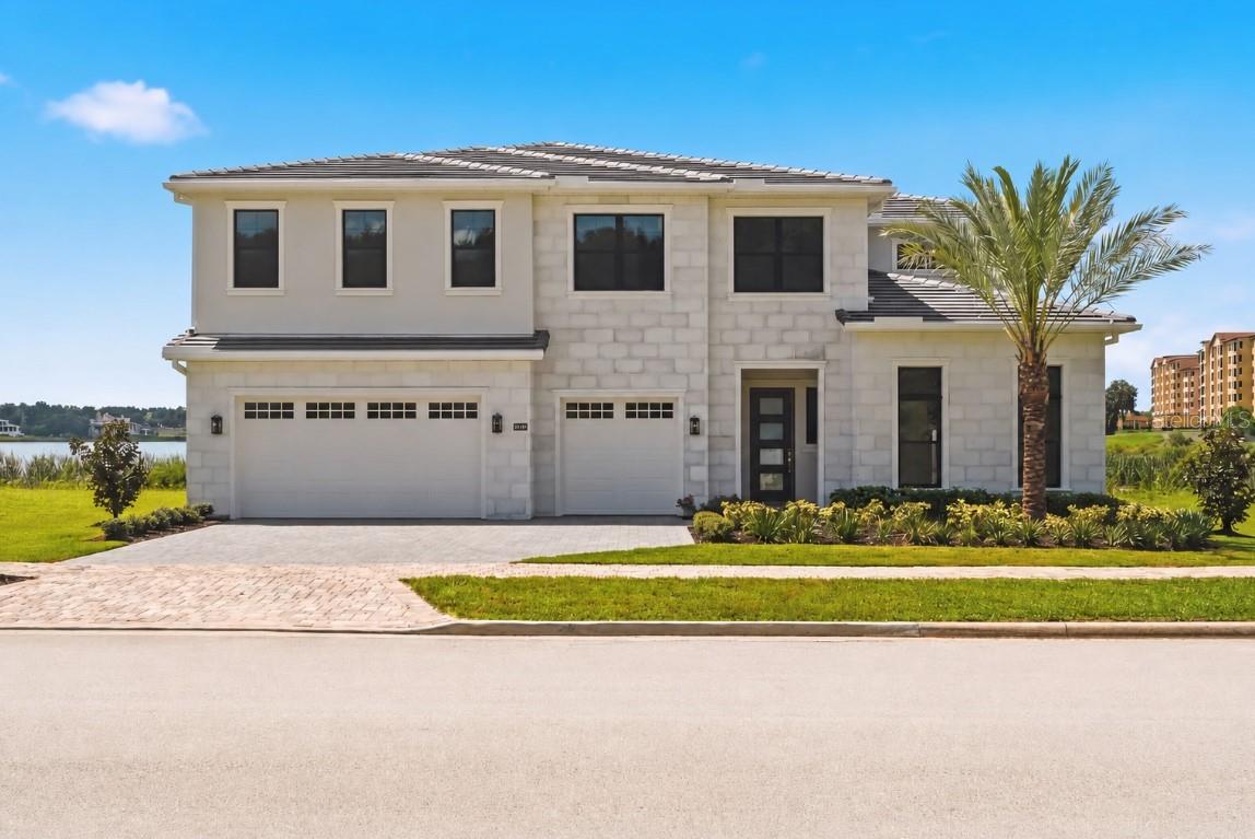 Two-story stone house with a double garage, palm tree, and lake visible behind it.