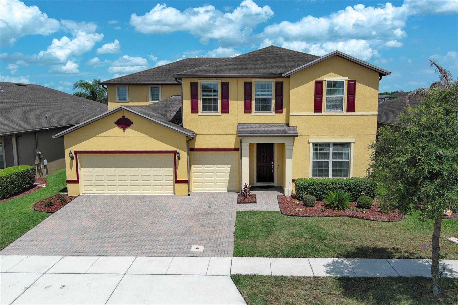 Two-story yellow house with burgundy shutters, a two-car garage, and a wide paver driveway.