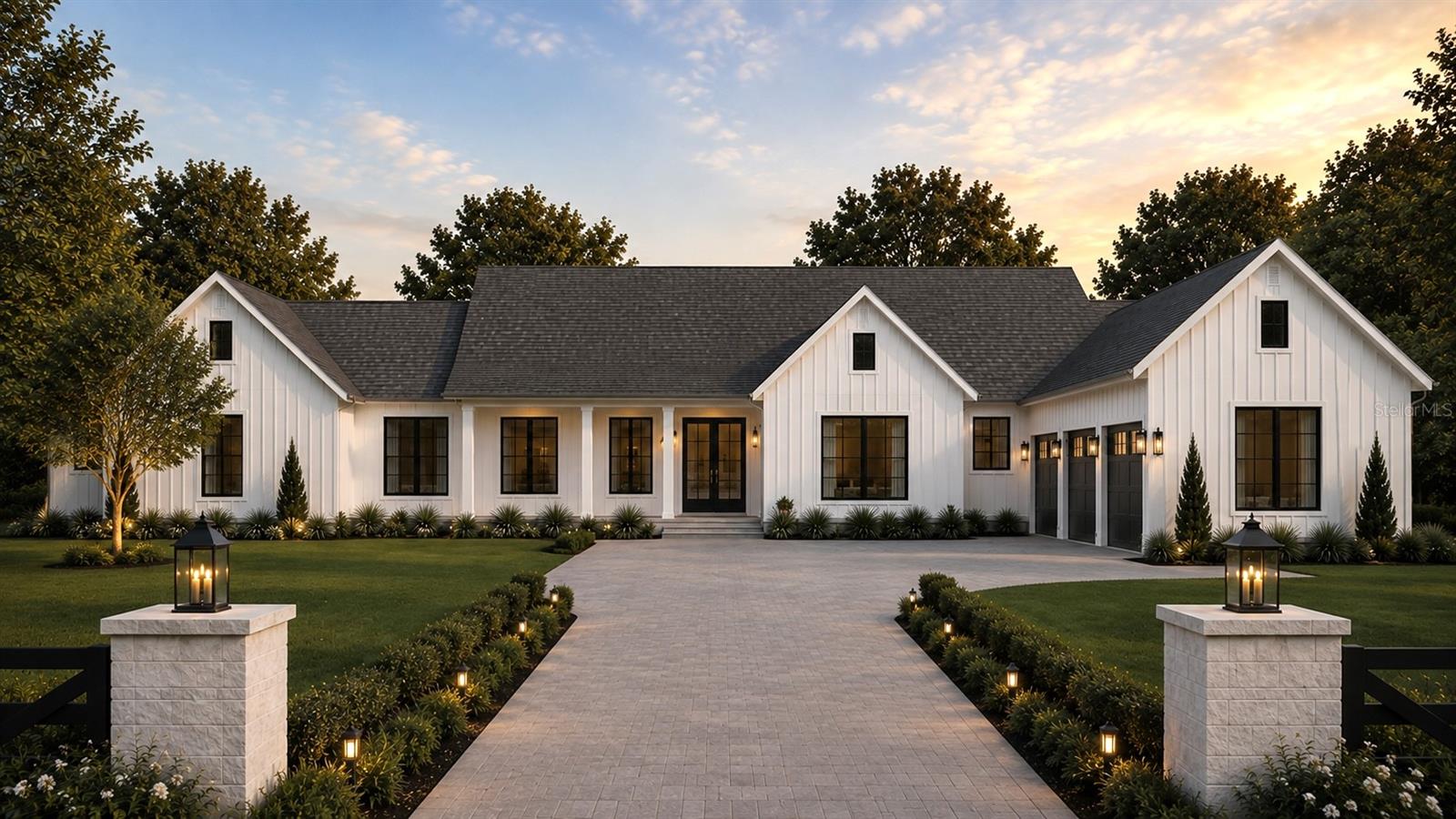 White modern farmhouse with black-framed windows, a broad paved driveway, and illuminated entrance pillars at dusk.