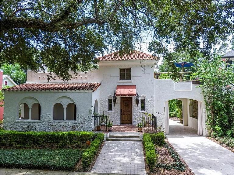 White stucco home with a red tile roof, arched windows, and a covered driveway beneath mature trees.