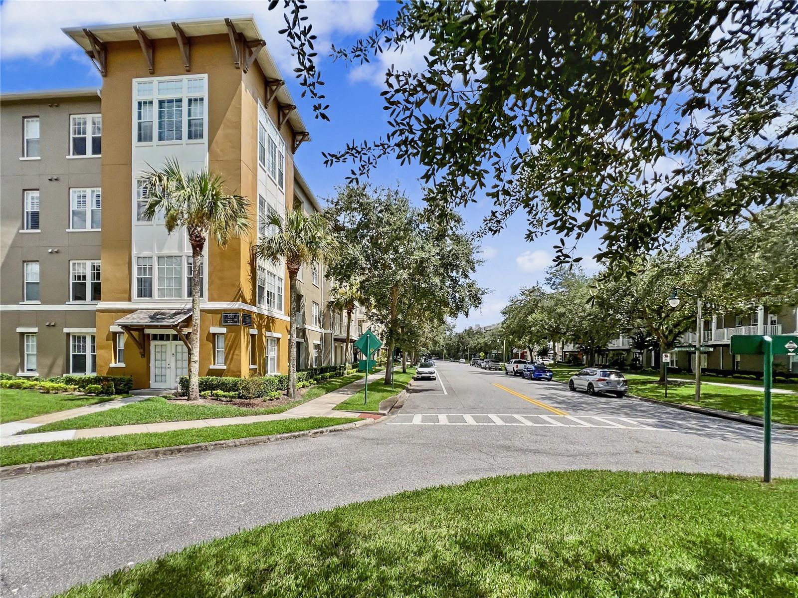 Tree-lined neighborhood street with a tan multi-story building, parked cars, and a crosswalk.