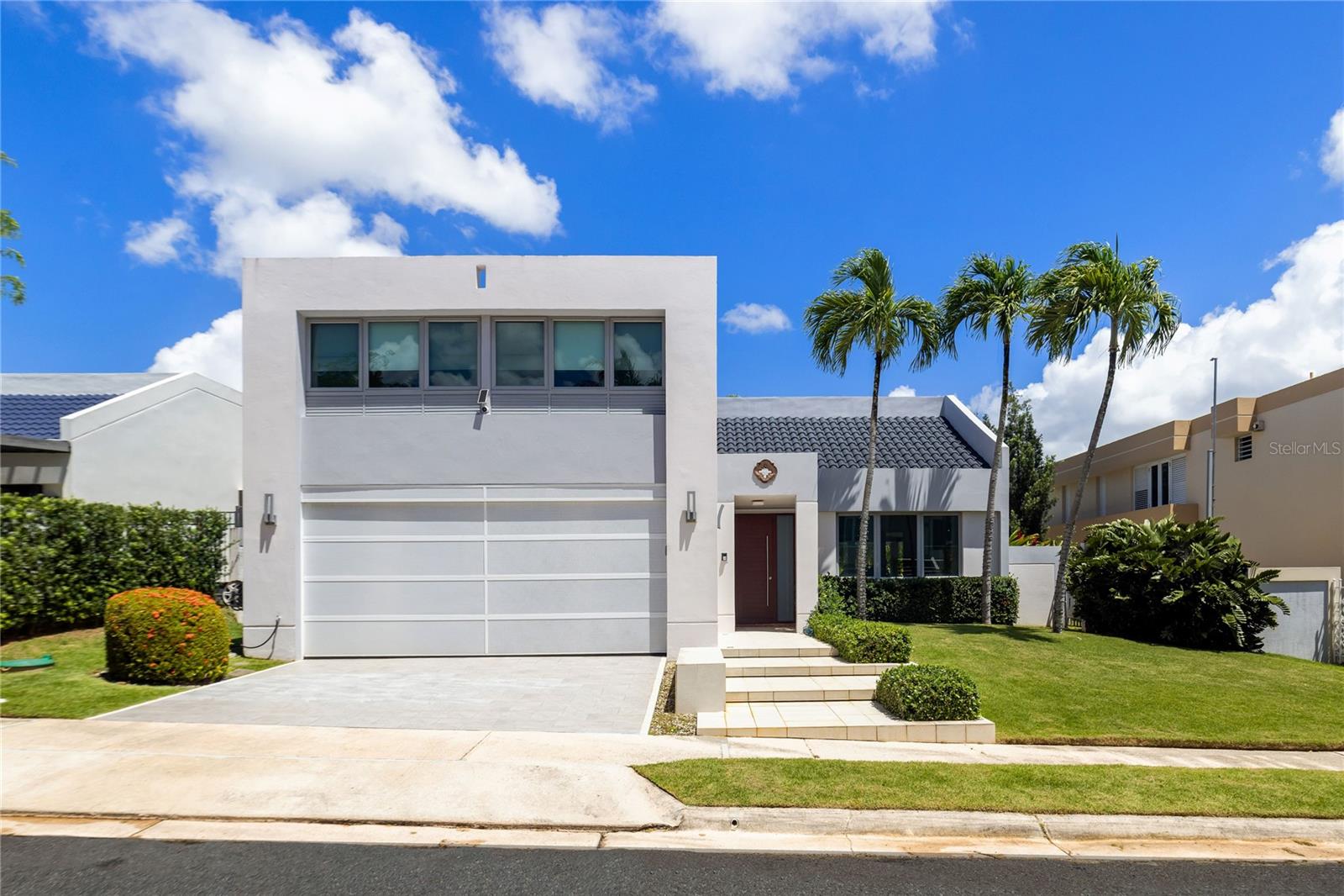 Modern white home with a large garage, landscaped lawn, and tall palm trees.