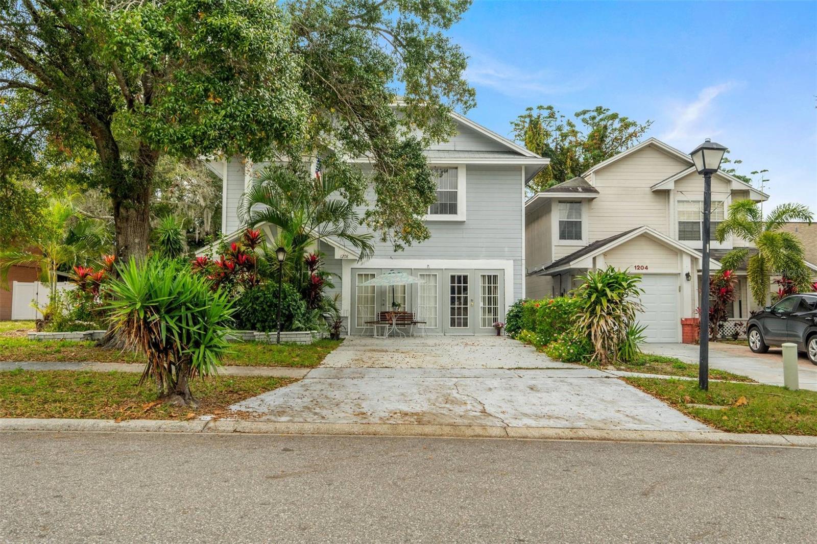 Light gray two-story home with glass patio doors, tropical landscaping, and a broad driveway.
