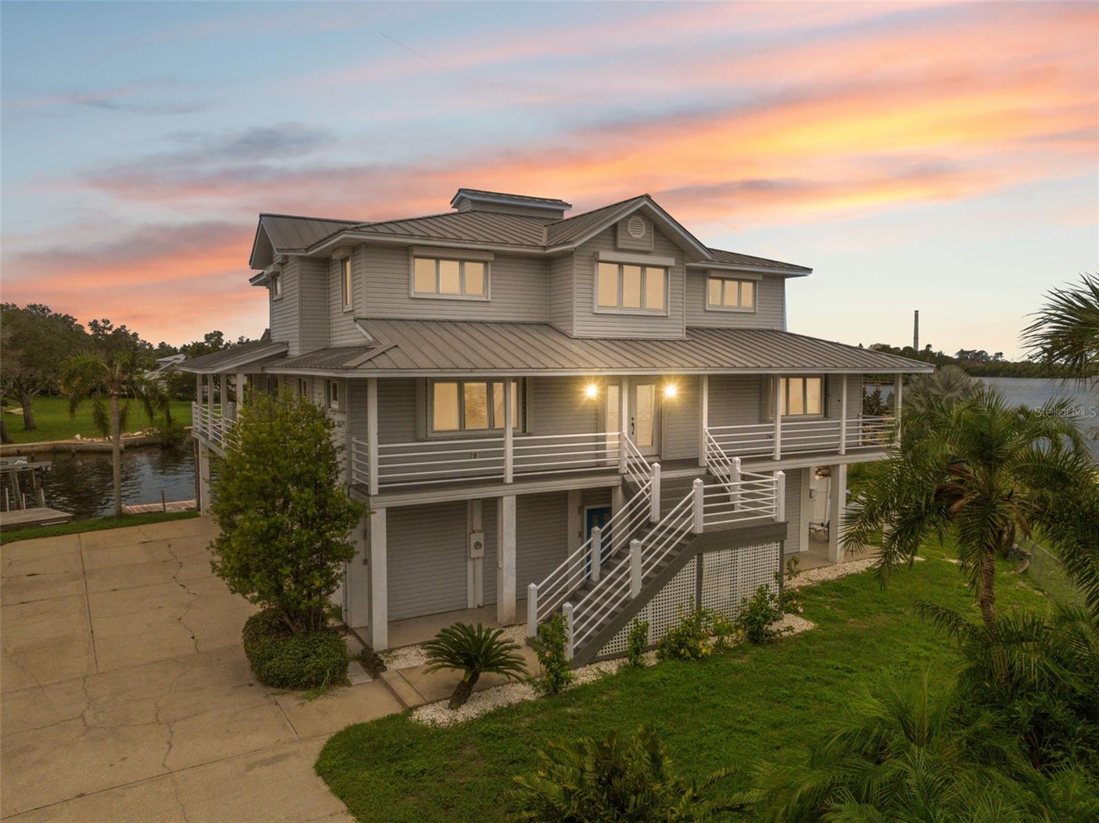 Elevated gray waterfront home with a wraparound porch beside a canal at sunset.