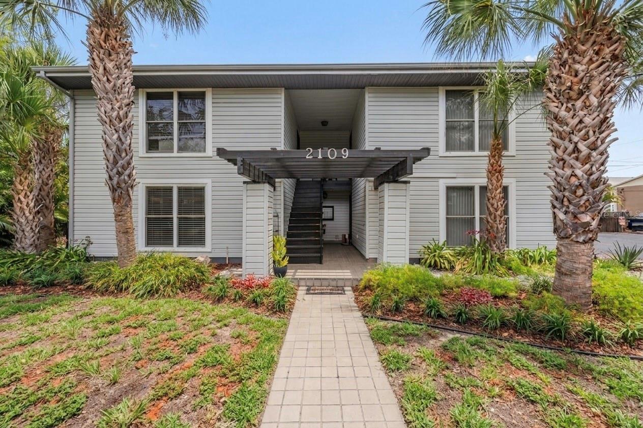Two-story multi-family building with a central exterior staircase, palm trees, and a landscaped front walkway.