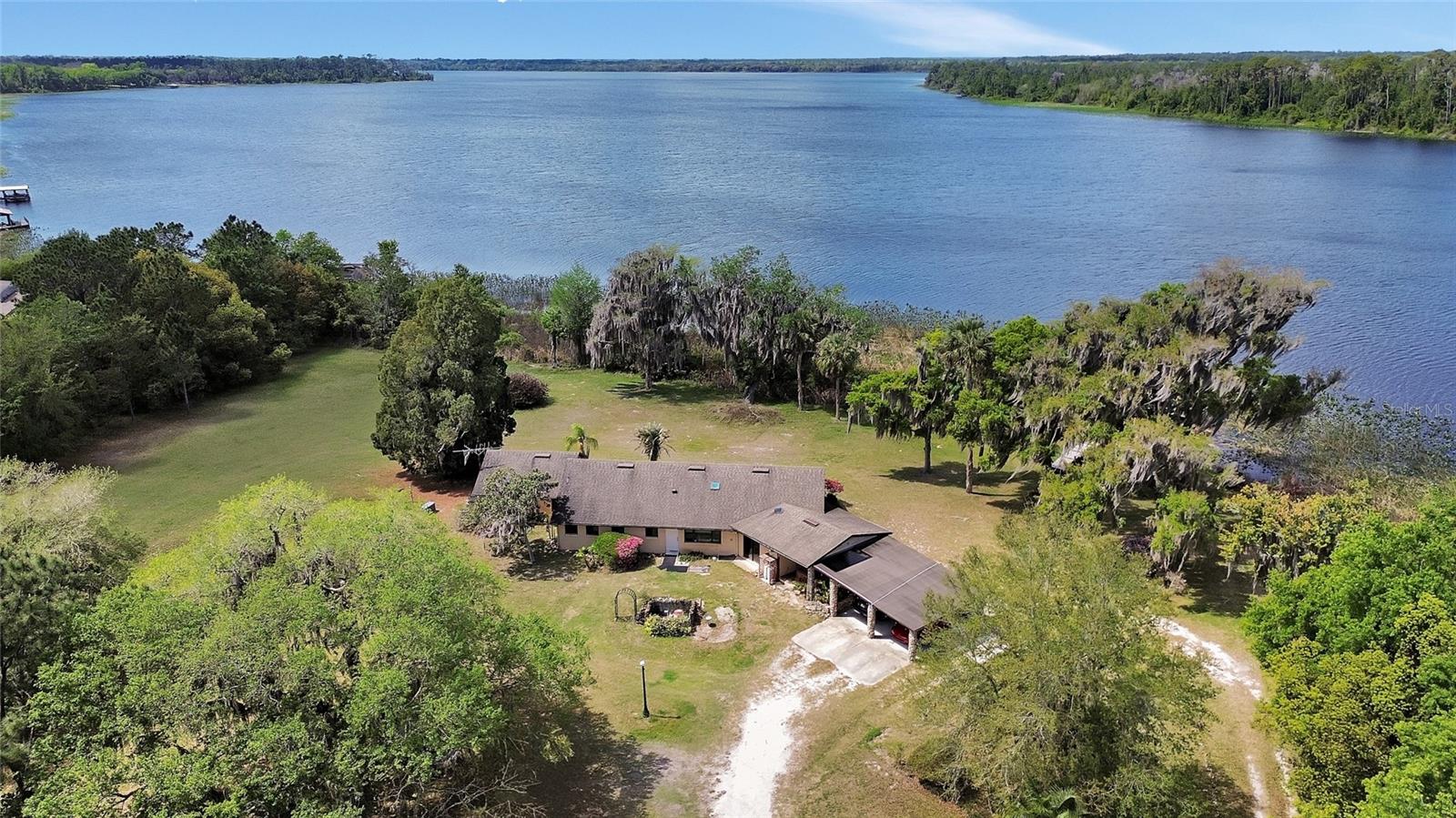 Aerial view of a lakeside home with a carport, open lawn, and mature trees.