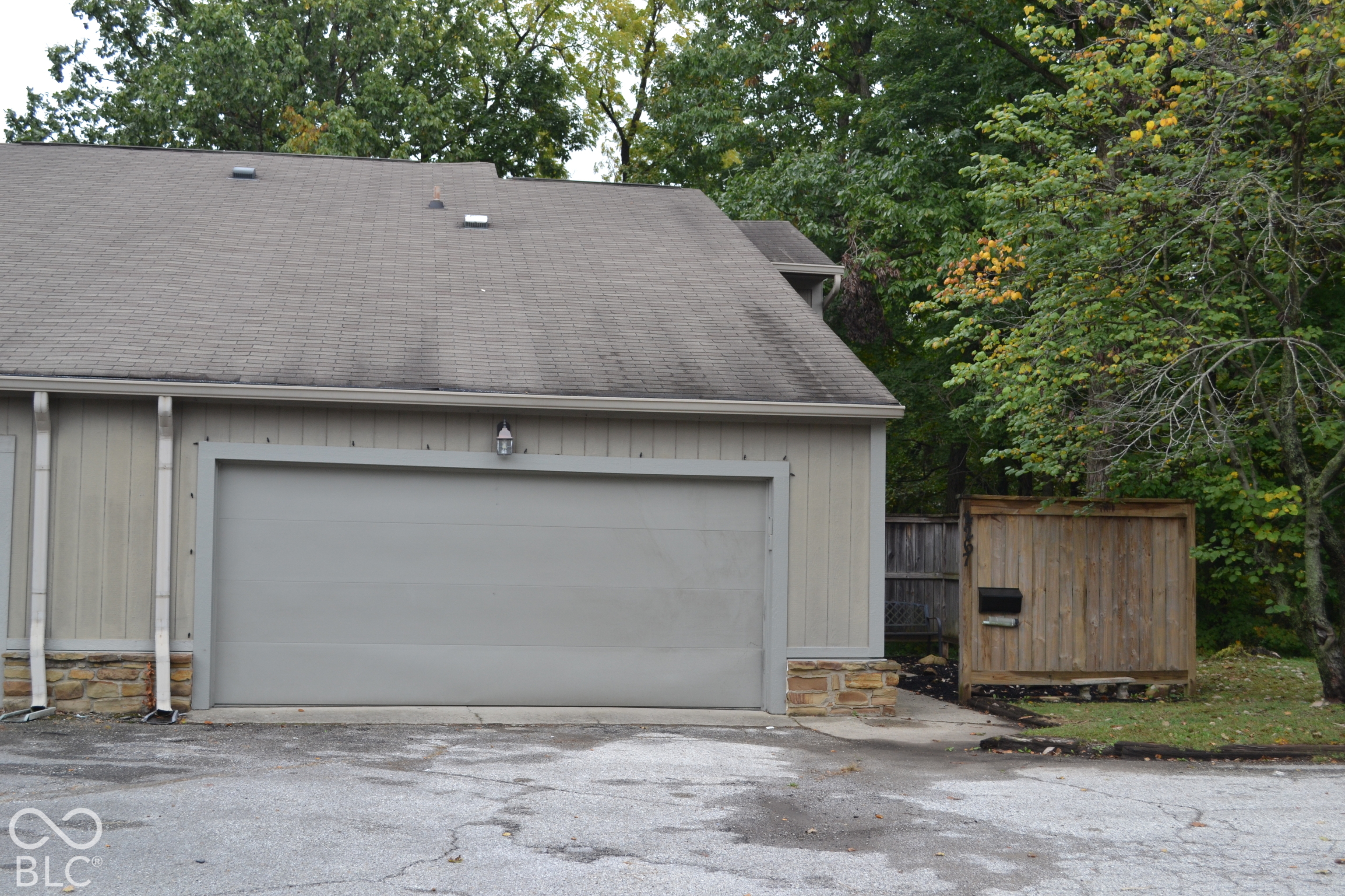 Gray detached garage with a large door, stone base, and wooded yard beside it.