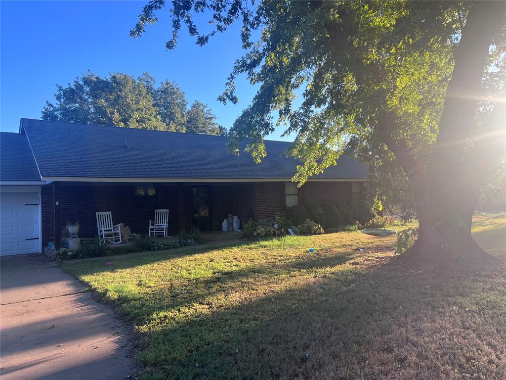 Brick ranch home with a covered porch, white rocking chairs, and a large shade tree.