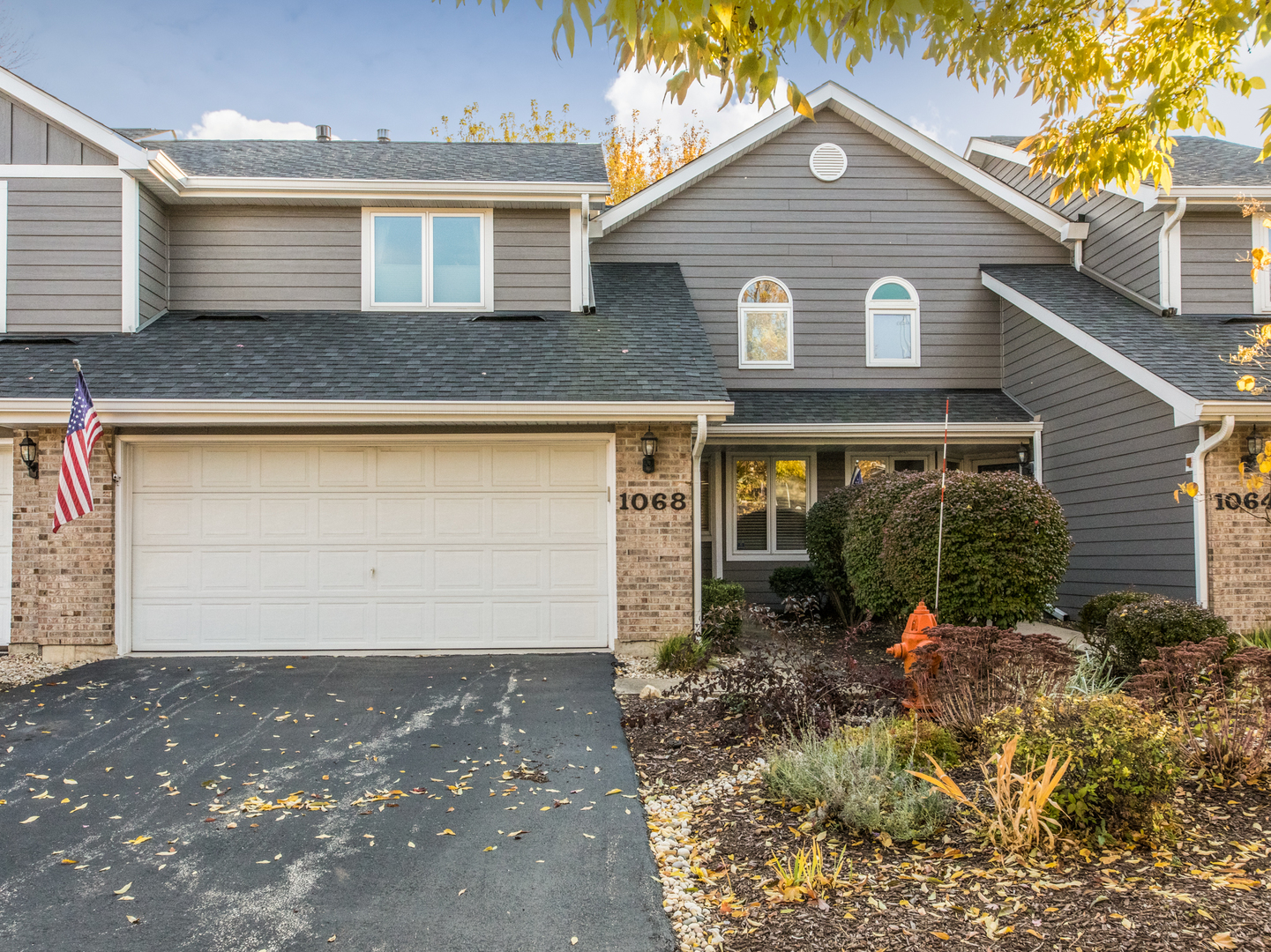 Gray two-story home with a white double garage, brick accents, landscaped garden, and American flag.
