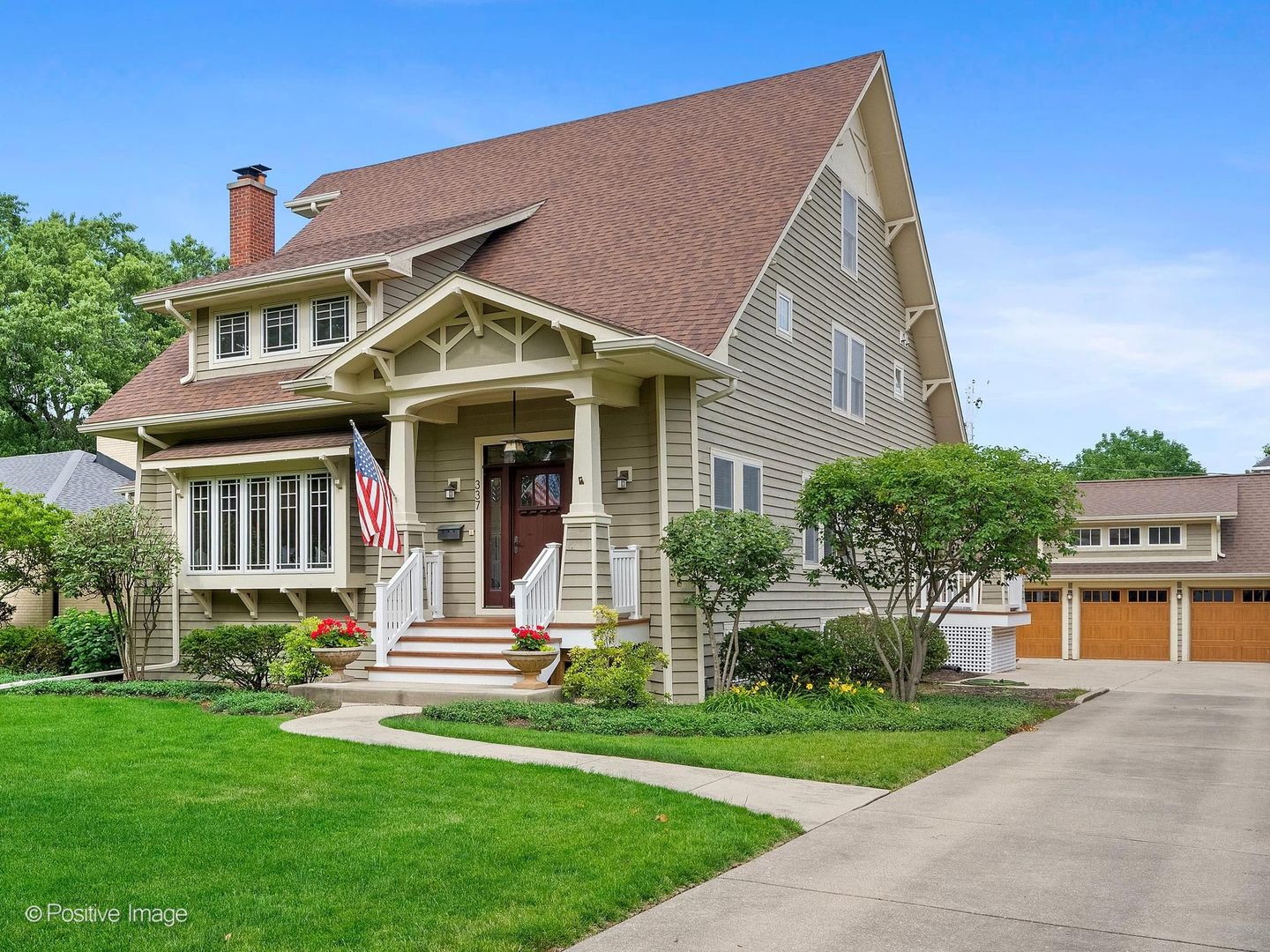Two-story gray-green house with a steep gabled roof, covered porch, American flag, and landscaped lawn.