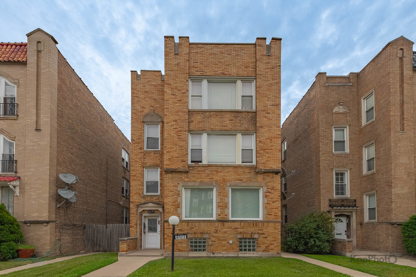 Three-story brick apartment building with a small front lawn, flanked by neighboring brick buildings.