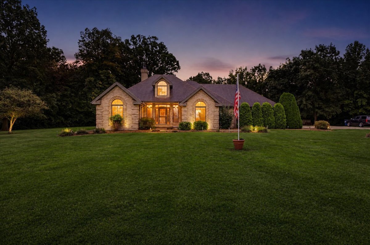 Stone-front home with warmly lit windows, a broad lawn, and an American flag at dusk.